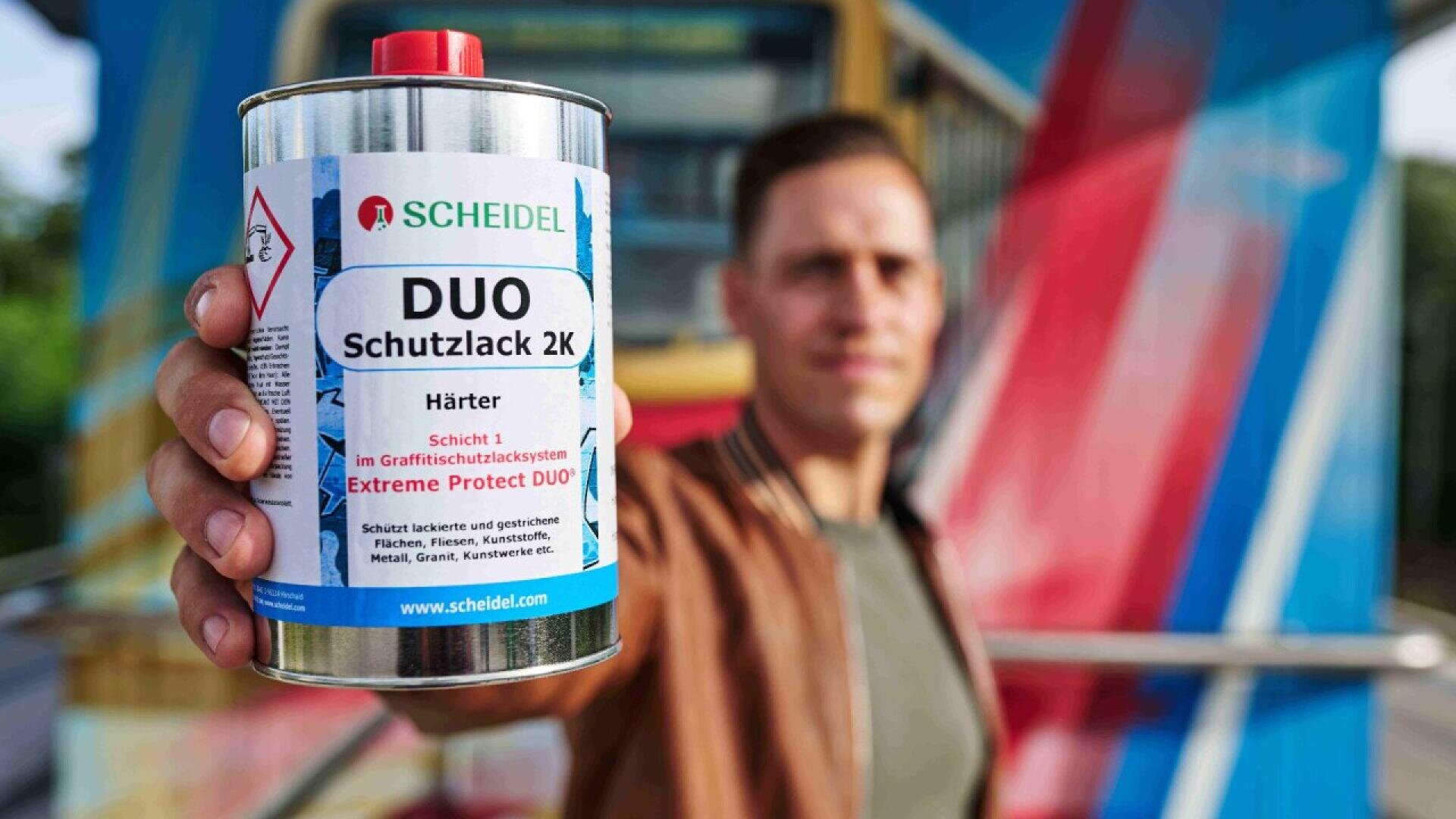 A person holds a can of Scheidel DUO Schutzlack 2K protective lacquer in front of a colorful graffiti-covered structure, with the product label clearly visible.