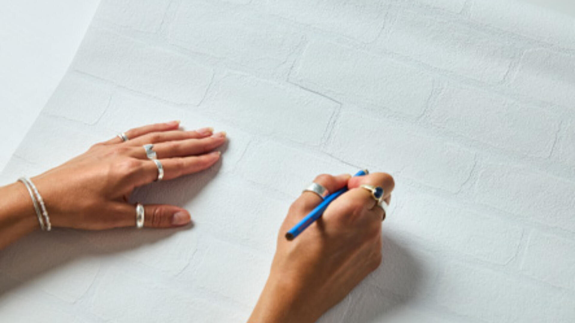 Eine Person, die mehrere Ringe und Armbänder trägt, zeichnet mit einem blauen Stift Ziegelmuster auf weißes Strukturpapier, das einer Ziegelmauer ähnelt.