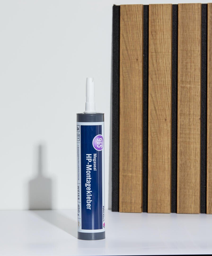 Eine Tube HP-Montagekleber steht aufrecht auf einer weißen Fläche neben einer Holzplatte mit vertikalen schwarzen Rillen.