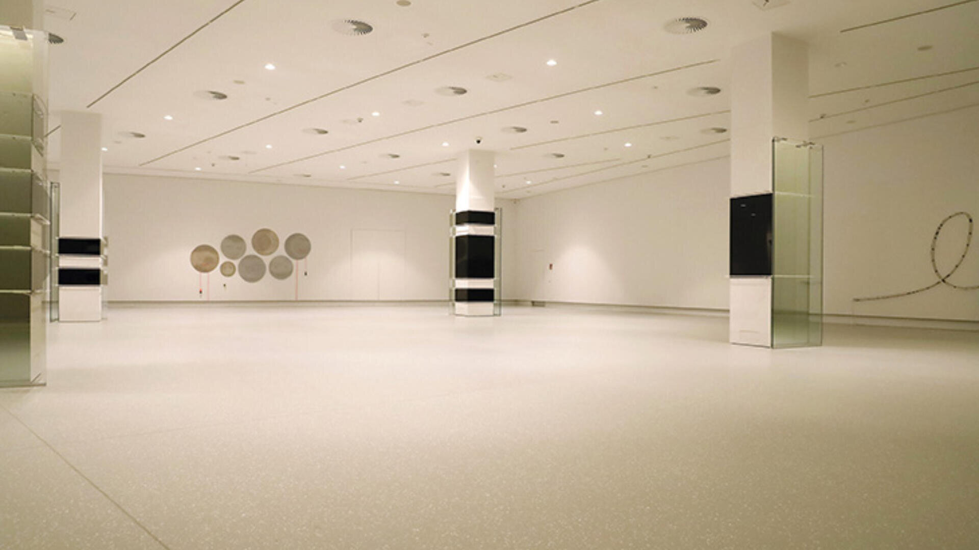 Eine geräumige, leere Kunstgalerie mit weißen Wänden, hellem Boden und mehreren Säulen. Minimalistische Wandkunst und Glasvitrinen sind im gesamten hellen Raum zu sehen.