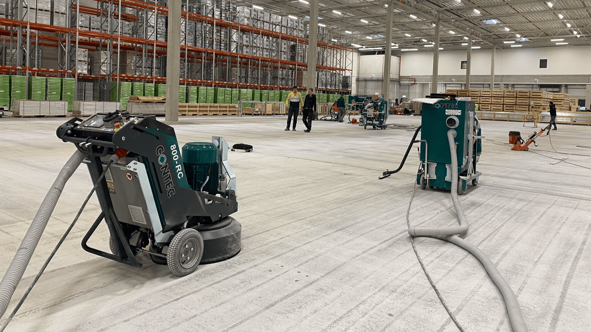 Industriemaschinen werden zum Reinigen oder Polieren des Betonbodens einer großen Lagerhalle mit hohen Decken und Regalen eingesetzt. Im Hintergrund sind einige Arbeiter in Sicherheitswesten zu sehen.