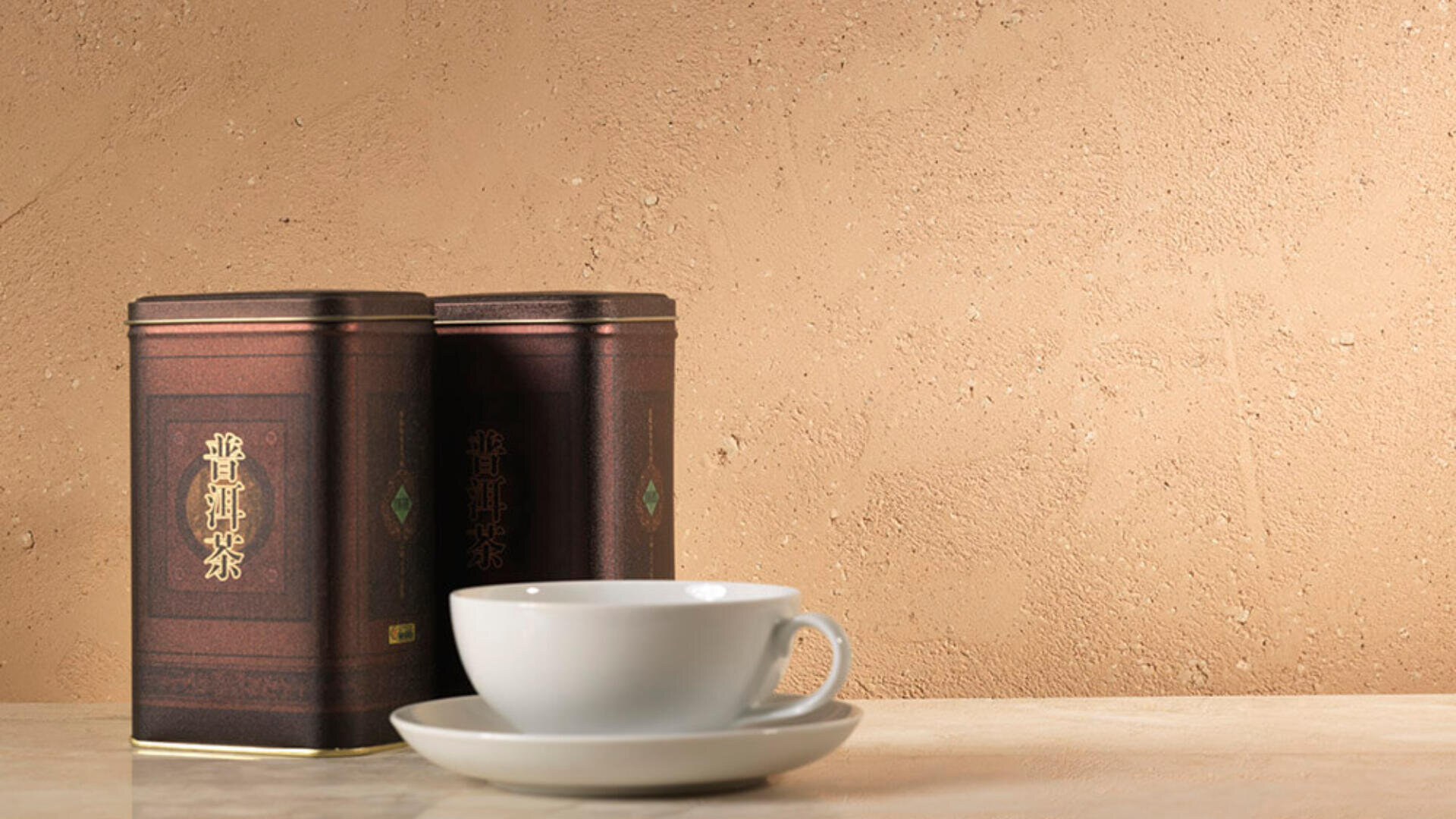 Eine weiße Teetasse und Untertasse stehen auf einer hellen Oberfläche neben zwei braunen Teedosen mit goldenen chinesischen Schriftzeichen vor einem strukturierten beigen Hintergrund.