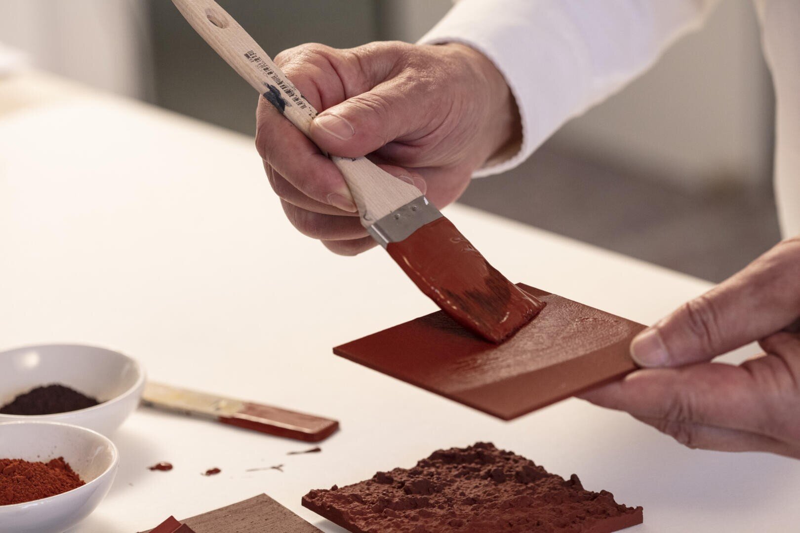 Eine Person trägt mit einem Pinsel ein rötlich-braunes Pigment auf eine kleine rechteckige Fliese auf. Auf dem Tisch sind Schüsseln mit Pigmentpulver und andere Fliesen zu sehen.