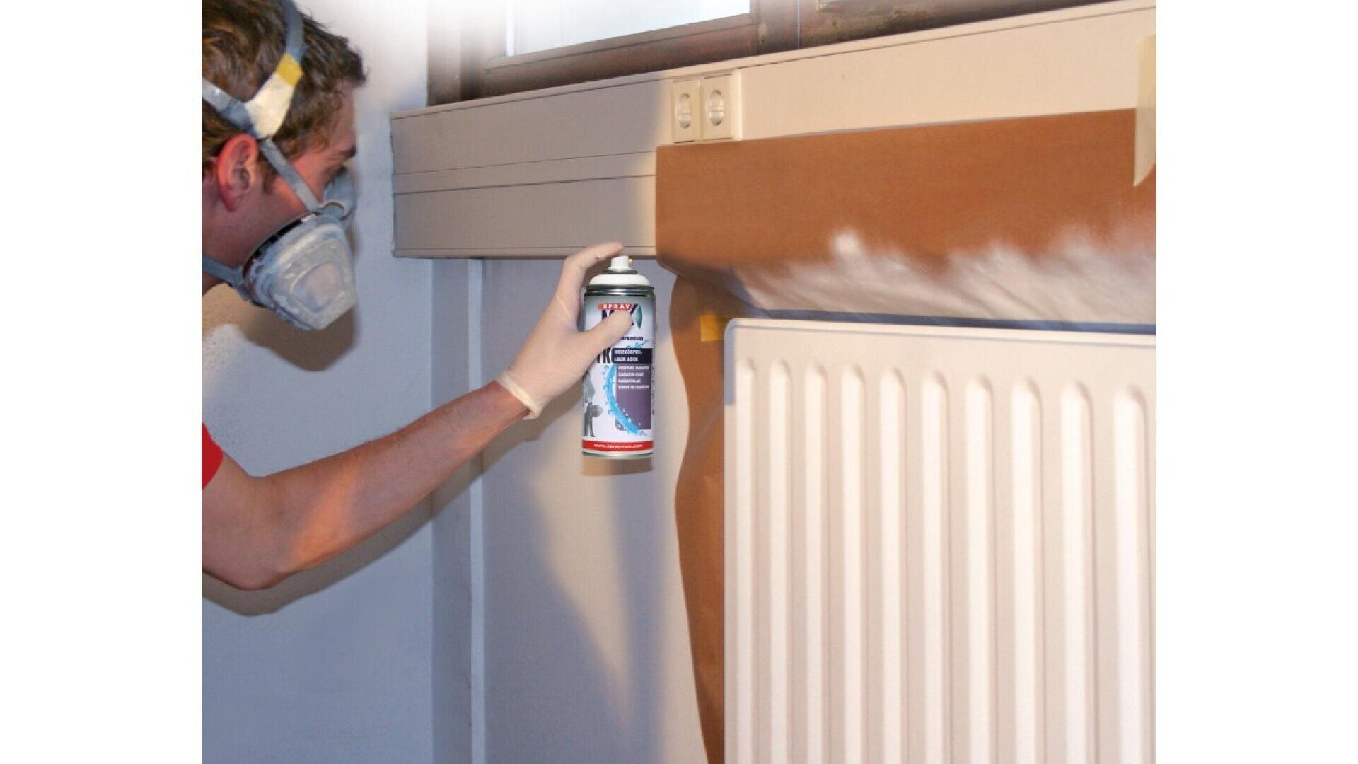 A person wearing a mask and gloves is spray painting a white radiator near a window. Brown paper protects the wall and window area from overspray.
