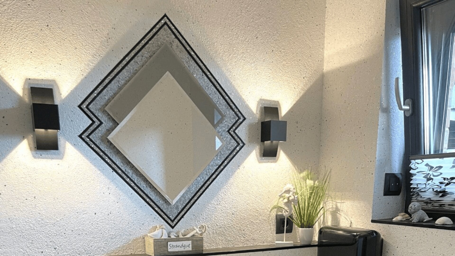 Ein modernes Badezimmer verfügt über einen rautenförmigen Spiegel, zwei Wandleuchten, eine kleine Pflanze auf einer Ablage und ein Fenster mit einem Milchglasmuster, das natürliches Licht hereinlässt.