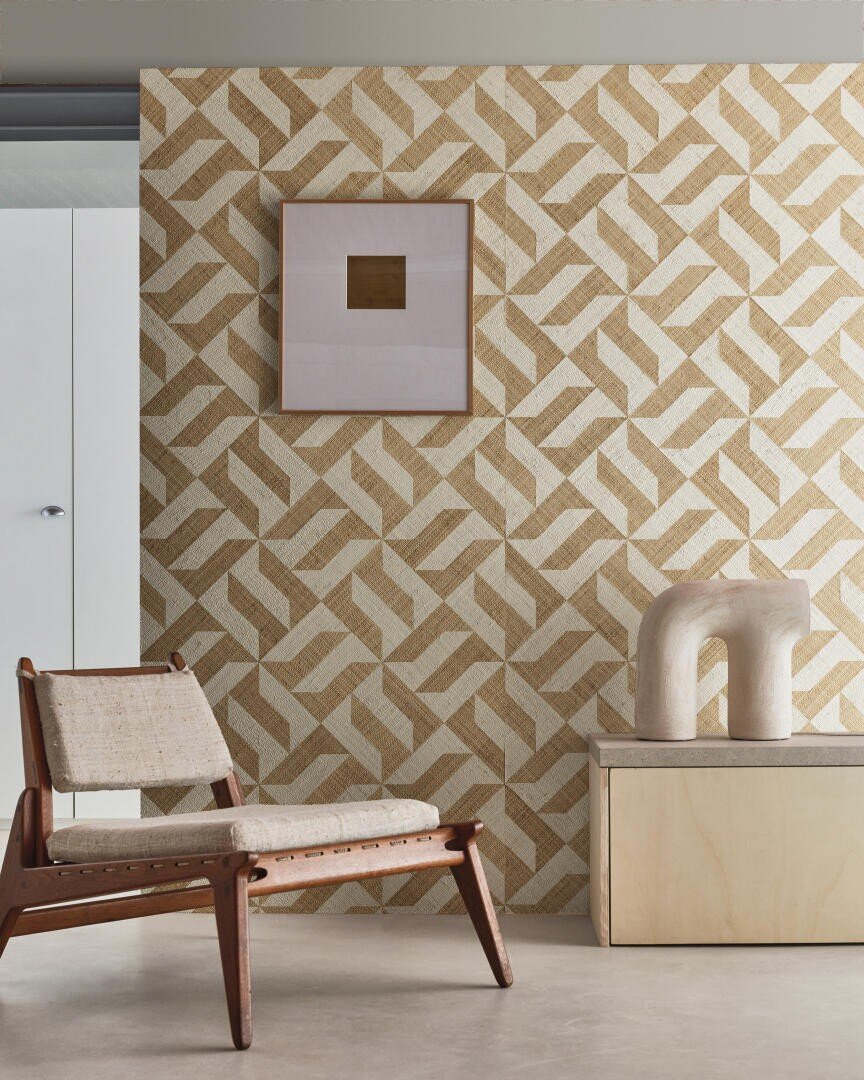 Ein modernes Interieur mit einer geometrisch gemusterten Wand in Beigetönen, einem geflochtenen Loungesessel, einem gerahmten minimalistischen Kunstwerk und einem skulpturalen Keramikstück auf einem hellen Holzschrank.