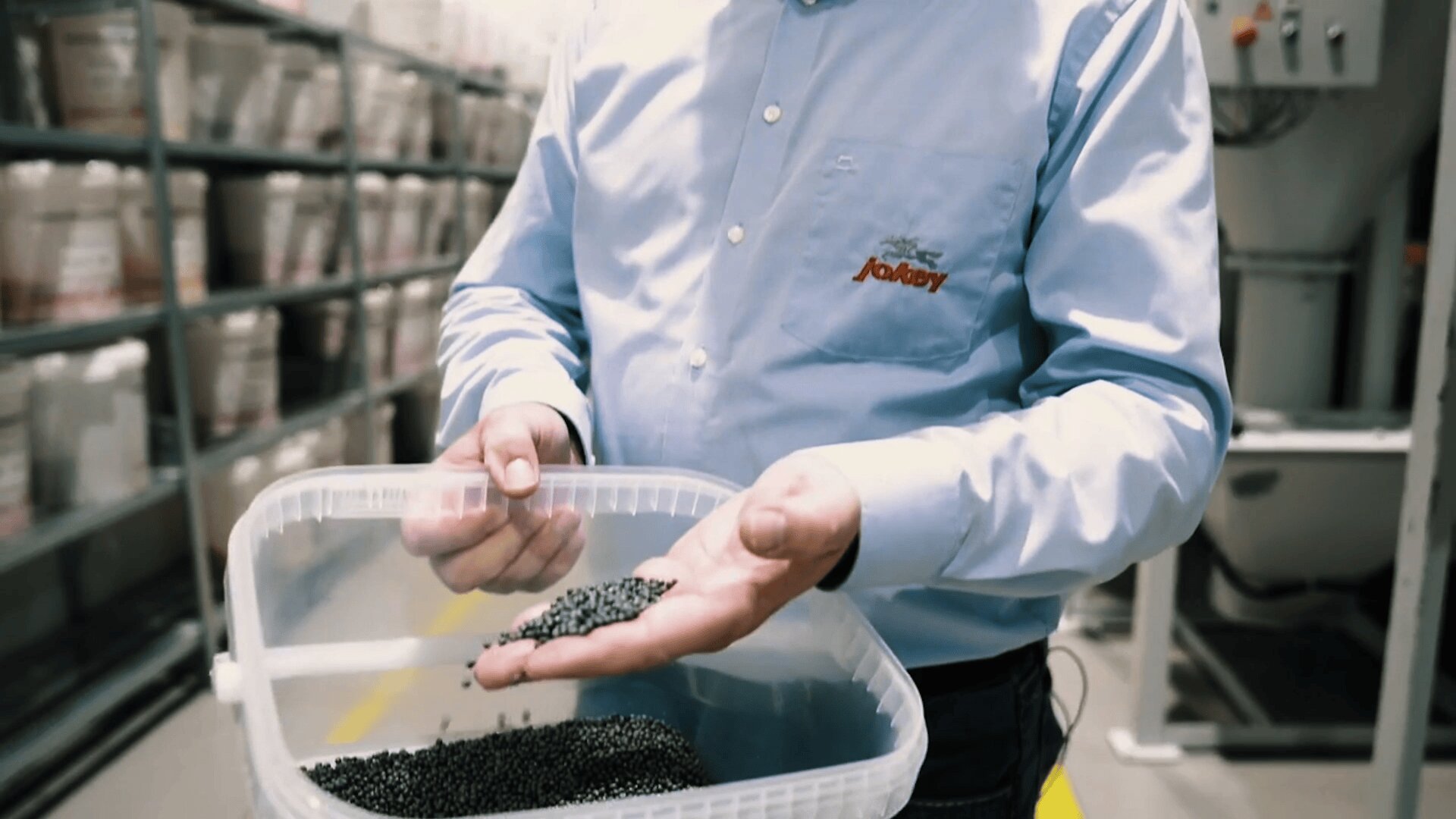 A person in a blue shirt stands in a warehouse, holding and inspecting small black plastic pellets from a clear container. Shelves with stacked containers are visible in the background.