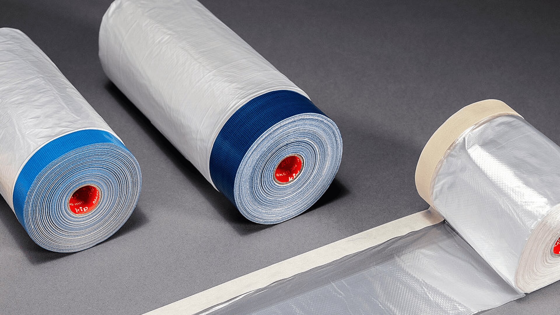 Drei Rollen transparenter Plastikfolie mit rotem Kern liegen aufgereiht auf einem dunklen Untergrund. Zwei Rollen haben blaue Bänder, die dritte hat ein cremefarbenes Band und ist teilweise abgerollt.