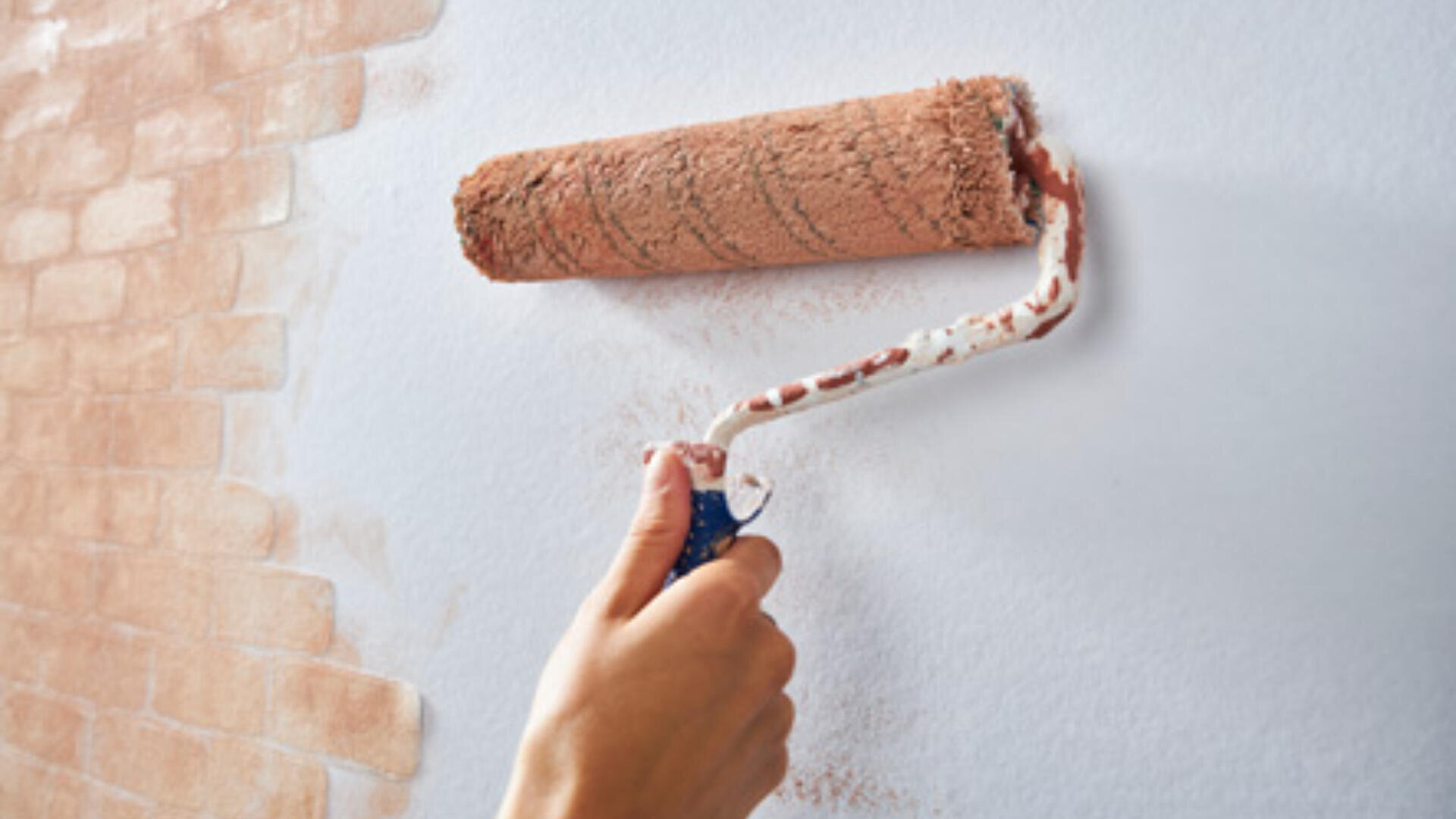 Eine Hand trägt mit einer Farbrolle eine hellbraune Farbe mit Ziegelmuster auf eine weiße Wand auf und erzeugt so einen künstlichen Ziegeleffekt.