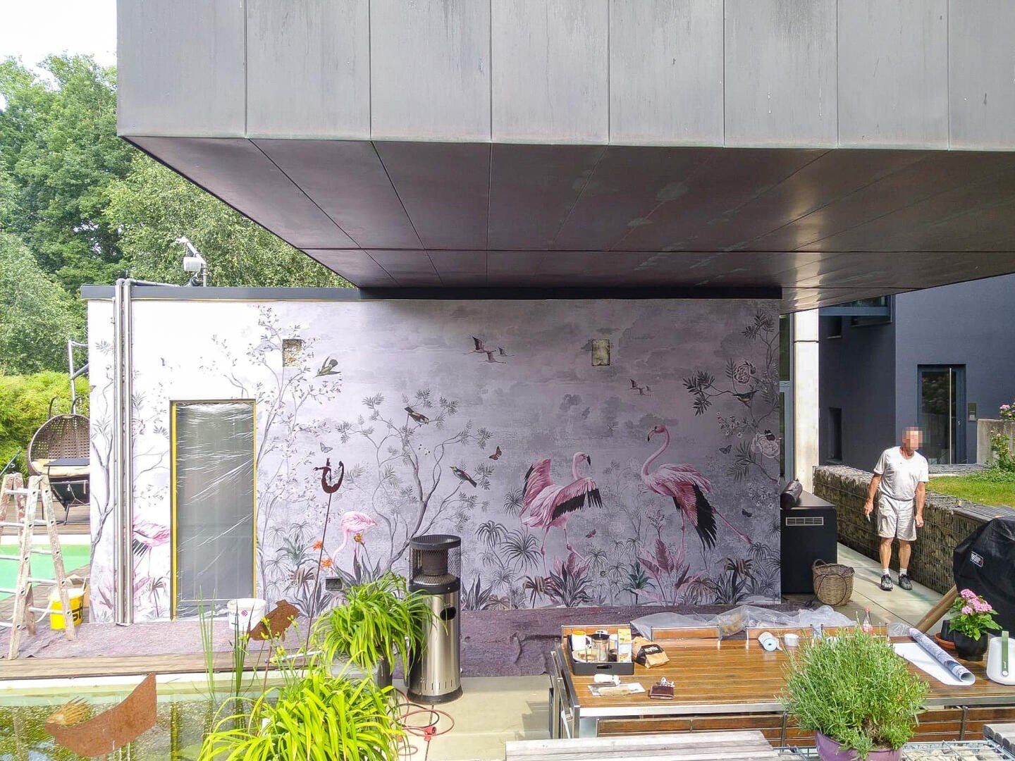 Eine moderne Terrasse mit einem Wandgemälde mit Flamingos, Pflanzen und Vögeln. Ein Mann steht neben einem Grill. Im Vordergrund stehen ein Holztisch und grüne Topfpflanzen. Das Haus besticht durch minimalistische Architektur und große Überhänge.