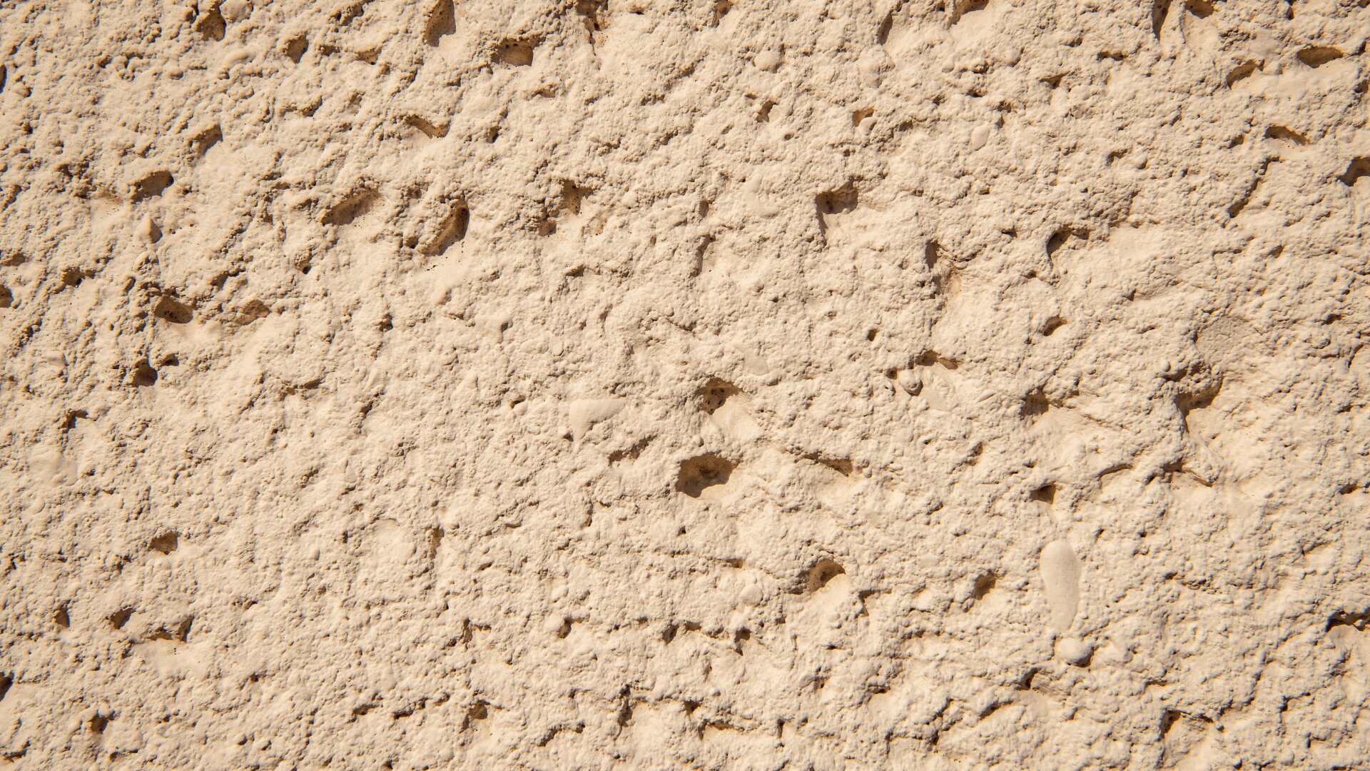 Nahaufnahme einer beigen, strukturierten Stuckwand mit unebener, rauer Oberfläche und kleinen Vertiefungen und Unebenheiten. Die Beleuchtung hebt die Textur und Tiefe des Stuckmaterials hervor.