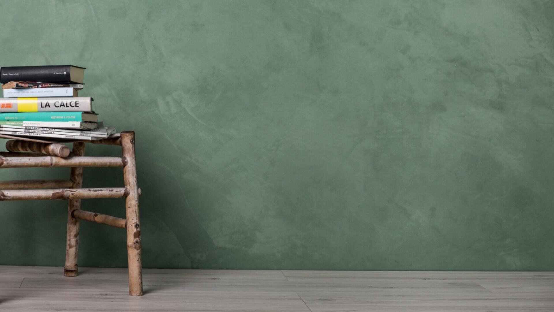 A stack of books sits on a rustic wooden stool against a green textured wall, with light gray wooden flooring beneath.
