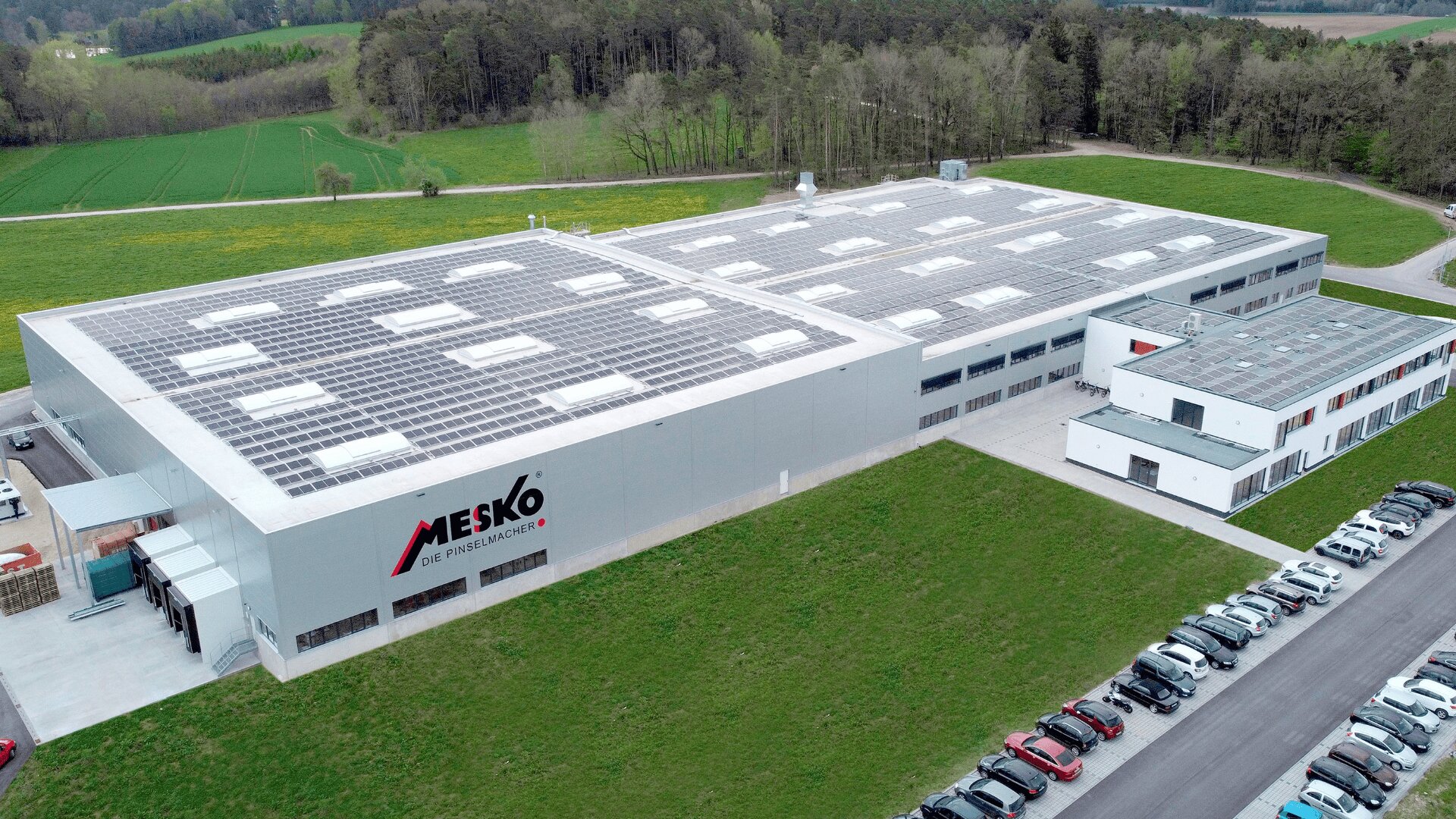 Luftaufnahme eines großen weißen Industriegebäudes mit dem MESKO-Logo an der Seite, Solarmodulen auf dem Dach, umgeben von Grün, einem Parkplatz mit Autos und einem nahegelegenen Wald.