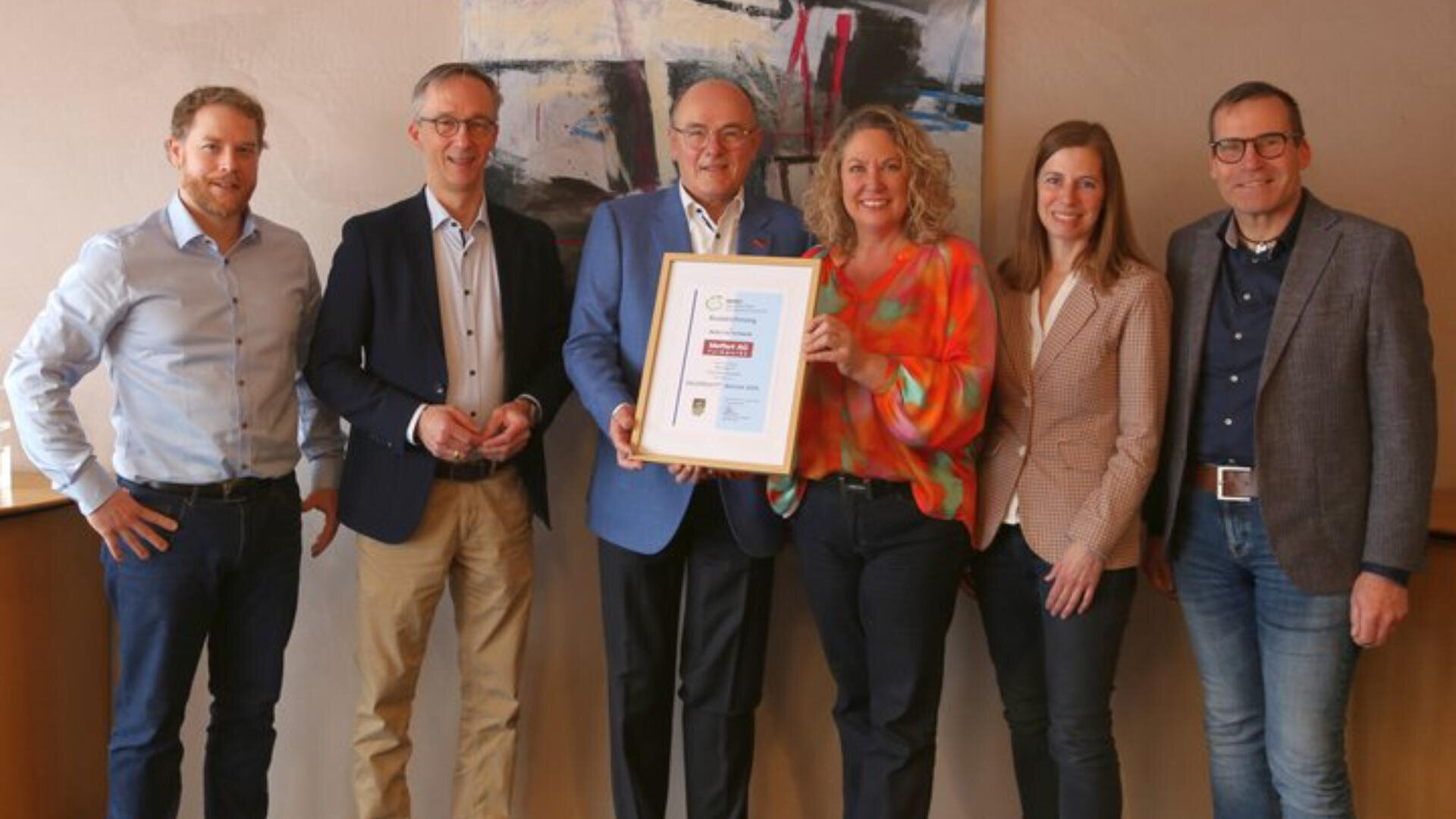 Sechs Personen stehen in einer Reihe im Innenbereich und lächeln in die Kamera. Die Frau in der Mitte hält eine gerahmte Urkunde oder Auszeichnung in den Händen. An der Wand dahinter hängt ein großes abstraktes Gemälde.