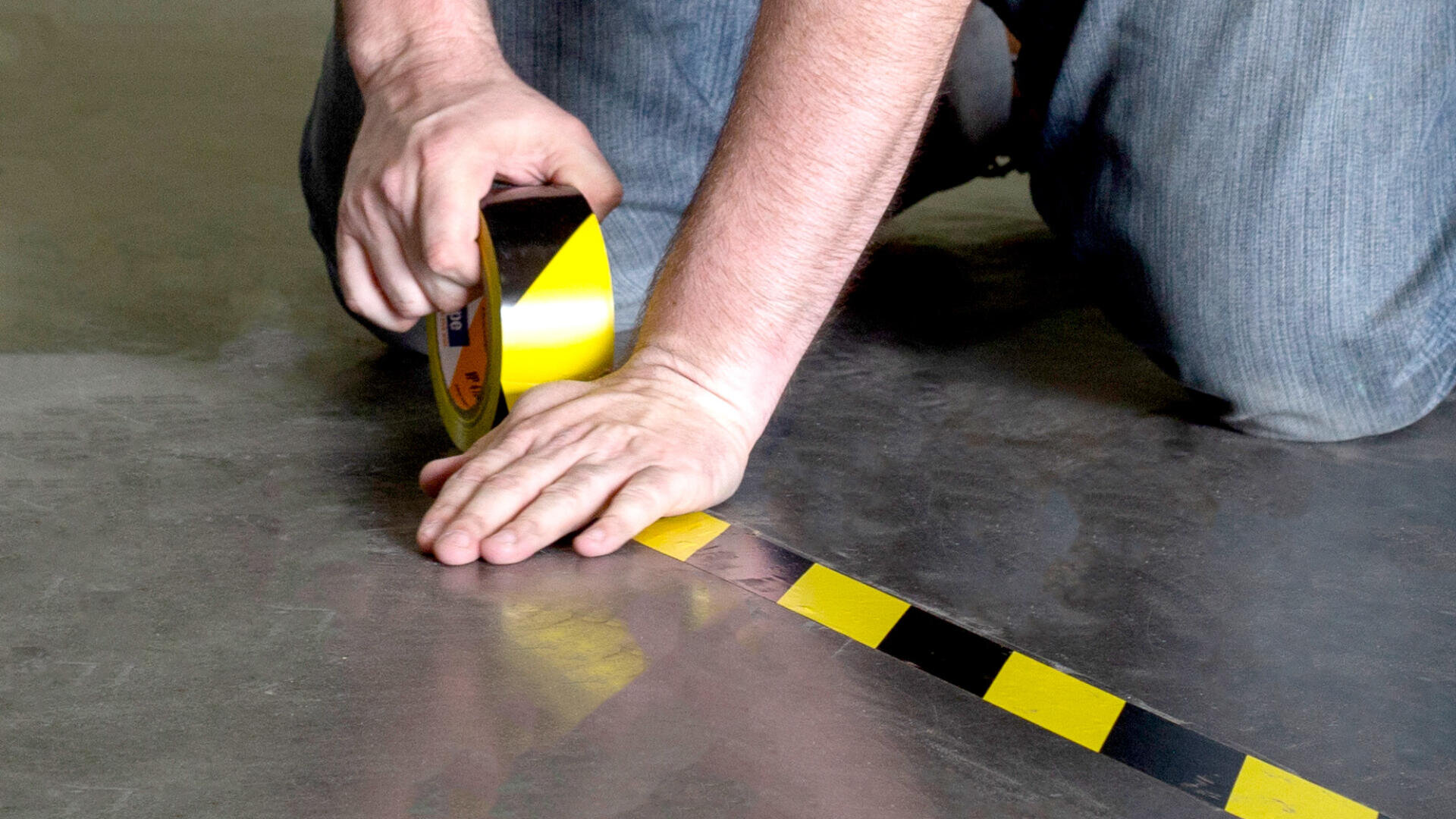 Eine auf dem Boden kniende Person bringt schwarz-gelb gestreiftes Sicherheitsklebeband auf einer glatten Oberfläche an, wobei sie mit einer Hand das Band nach unten drückt und mit der anderen die Rolle hält.