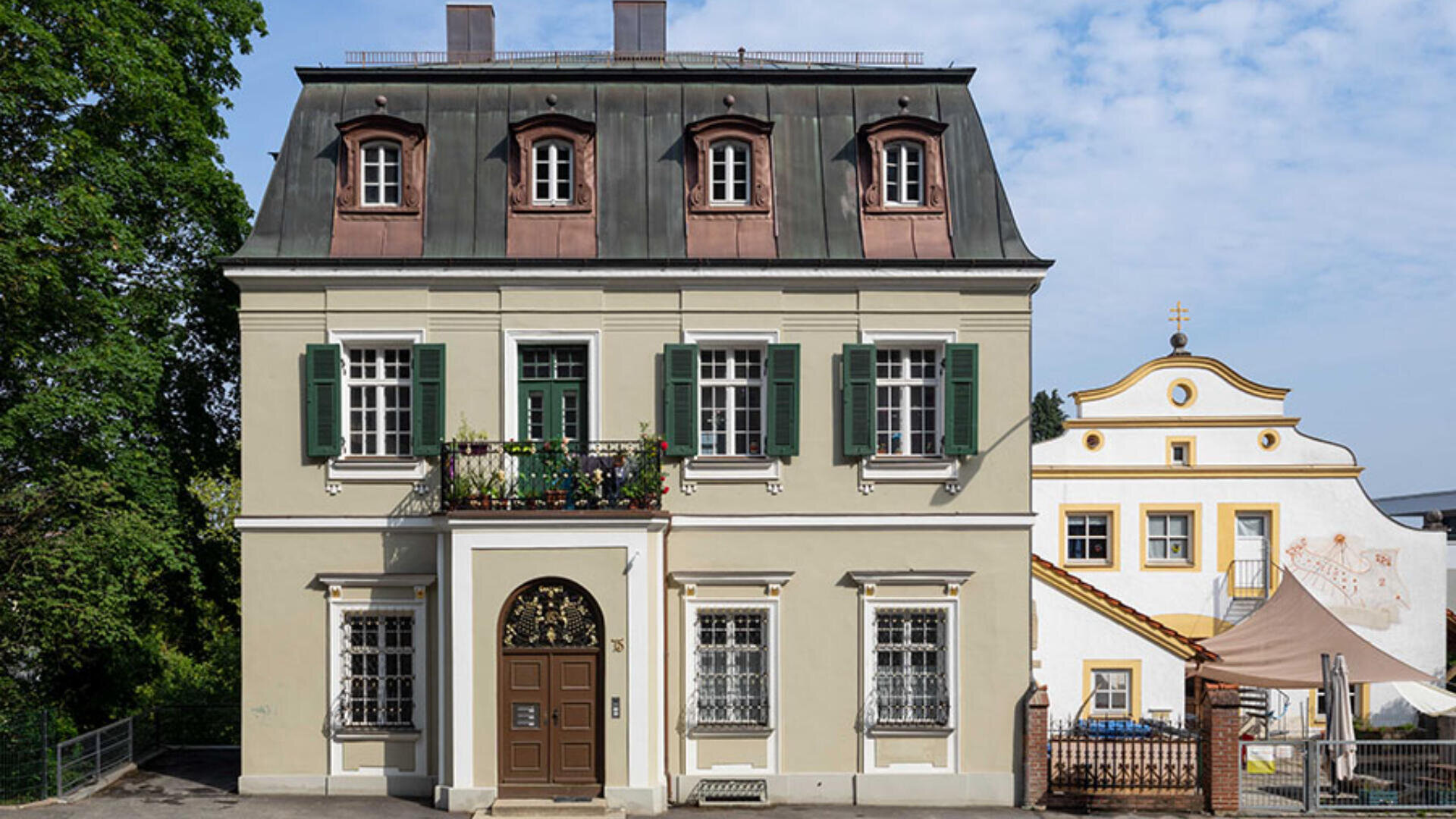 Ein zweistöckiges beiges Haus mit grünen Fensterläden und einem dekorativen Eisenbalkon ist vor einem weißen Gebäude mit einem geschwungenen Giebel und einem goldenen Kreuz darauf unter einem teilweise bewölkten Himmel zu sehen.