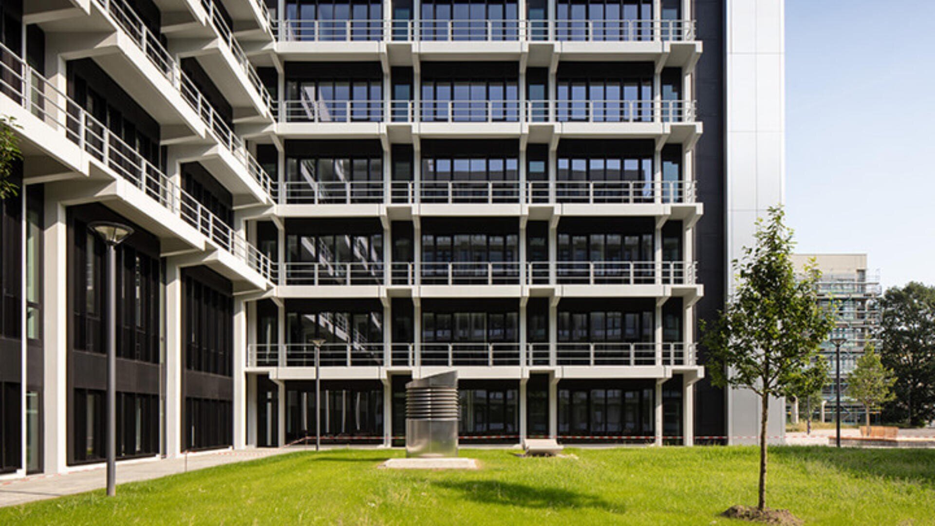 Modernes mehrstöckiges Gebäude mit großen Fenstern und Balkonen, mit Blick auf einen grünen Rasen mit einigen jungen Bäumen und einer kleinen Metallskulptur in der Mitte.