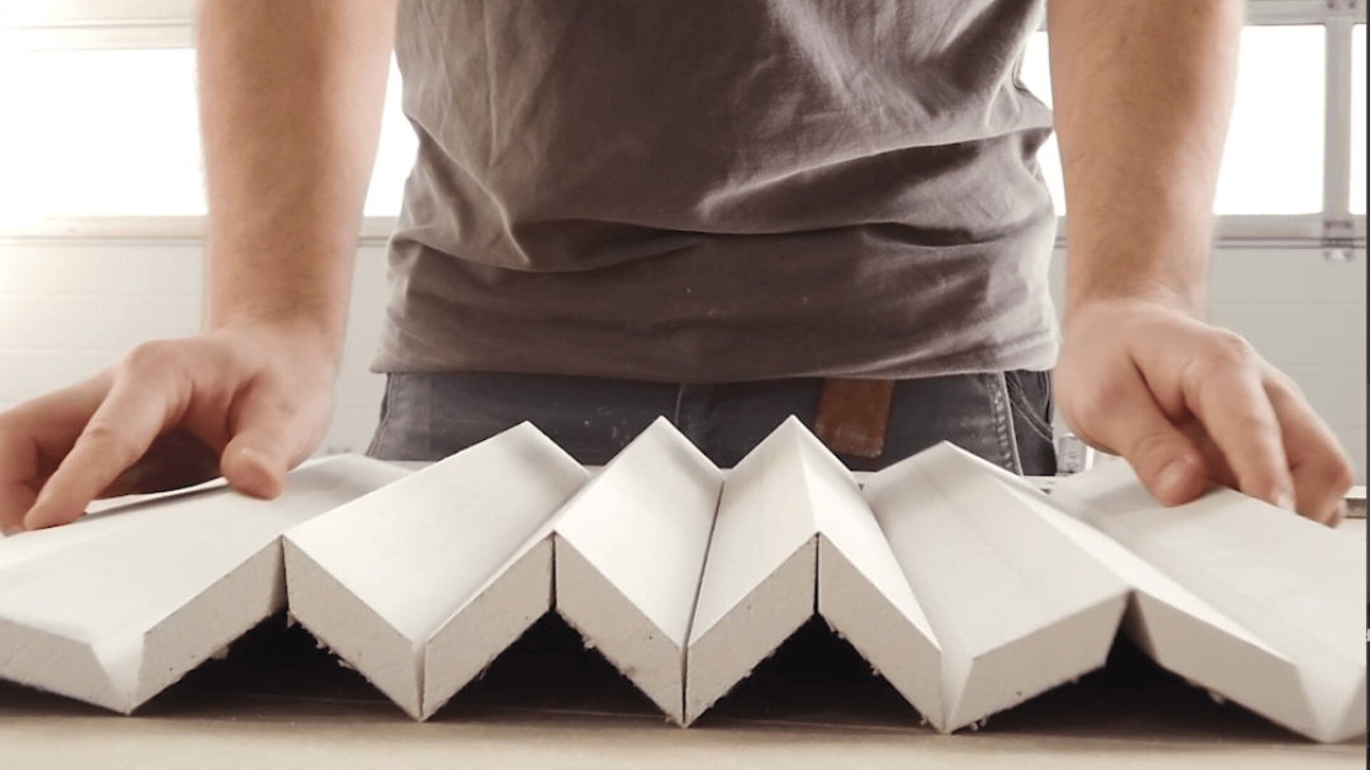 Eine Person in einem grauen Hemd ordnet zickzackförmige weiße Paneele auf einer flachen Oberfläche an, von der Hüfthöhe aus betrachtet.