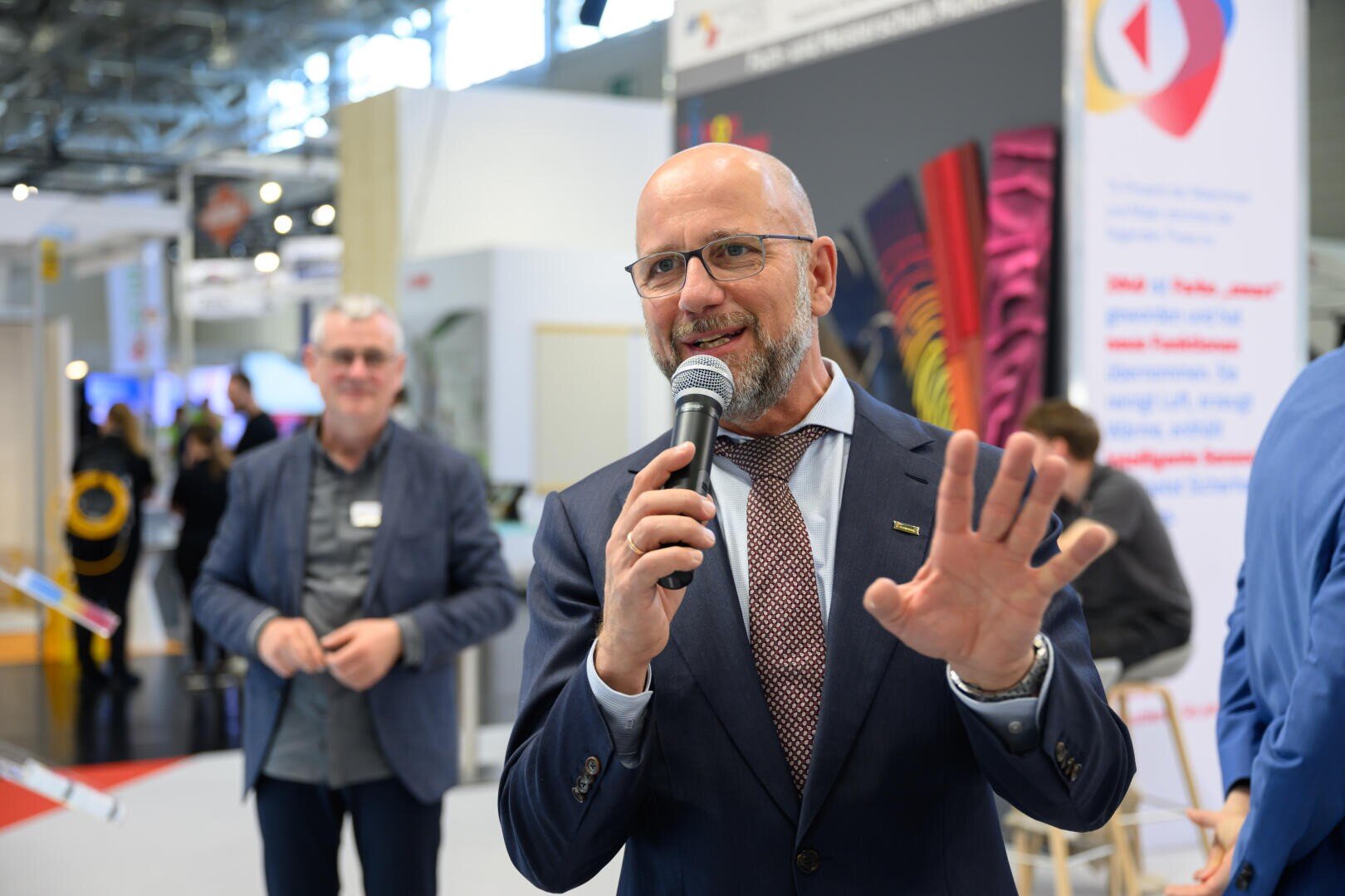 Ein glatzköpfiger Mann in Anzug und Brille spricht bei einer Indoor-Veranstaltung in ein Mikrofon und gestikuliert mit der Hand. Im Hintergrund sind weitere Personen und Messestände zu sehen.