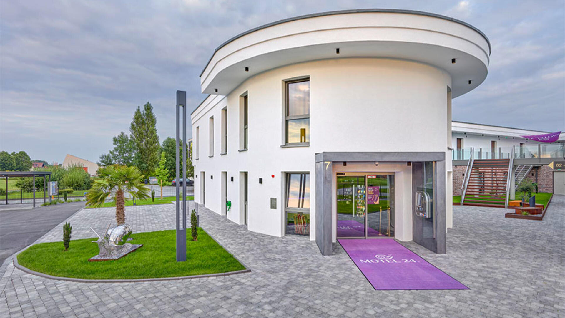 Ein modernes zweistöckiges Hotelgebäude mit großen Fenstern und geschwungenem Design, umgeben von gepflegten Rasenflächen, Palmen und einer gepflasterten Auffahrt. Auf einer lila Fußmatte am Eingang steht „Motel 24“.