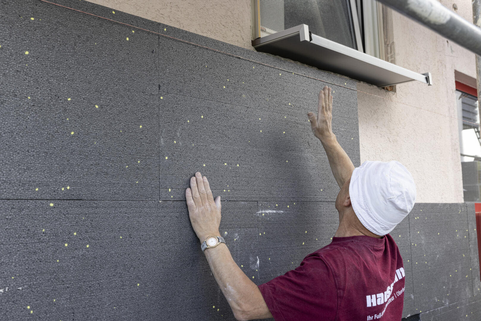 Ein Arbeiter mit einer weißen Kopfbedeckung installiert Dämmplatten an der Außenwand eines Gebäudes und greift nach oben, um eine Platte unter einem Fenster zurechtzurücken.