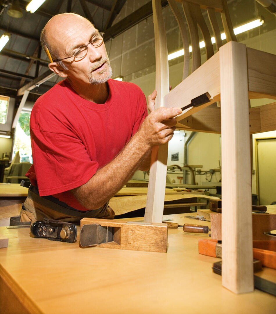 Ein Mann in einem roten Hemd bearbeitet in einer Schreinerei vorsichtig mit einem Meißel einen Holzstuhl, umgeben von Holzbearbeitungswerkzeugen auf einem Tisch.