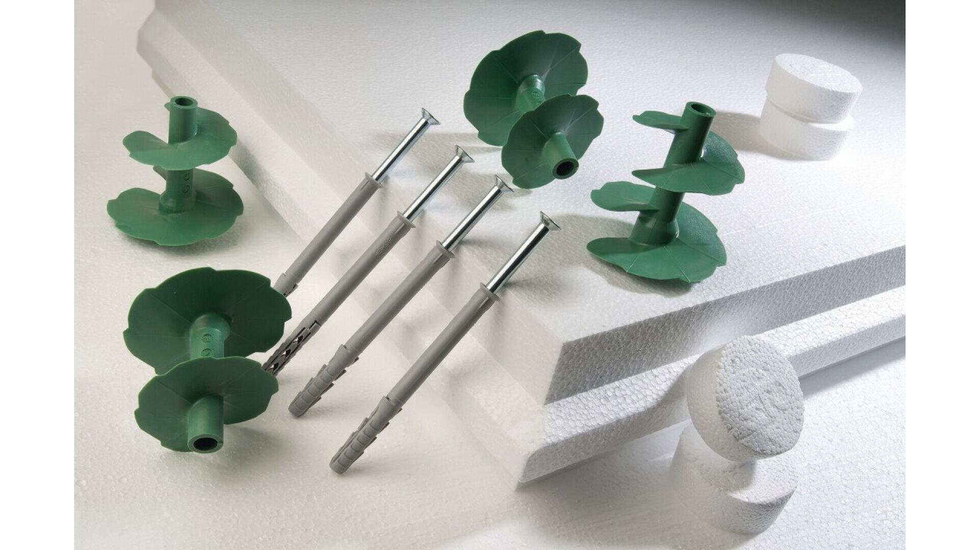 Four green plastic screw anchors and four metal screws are placed on large sheets and round pieces of white styrofoam, arranged on a flat surface.