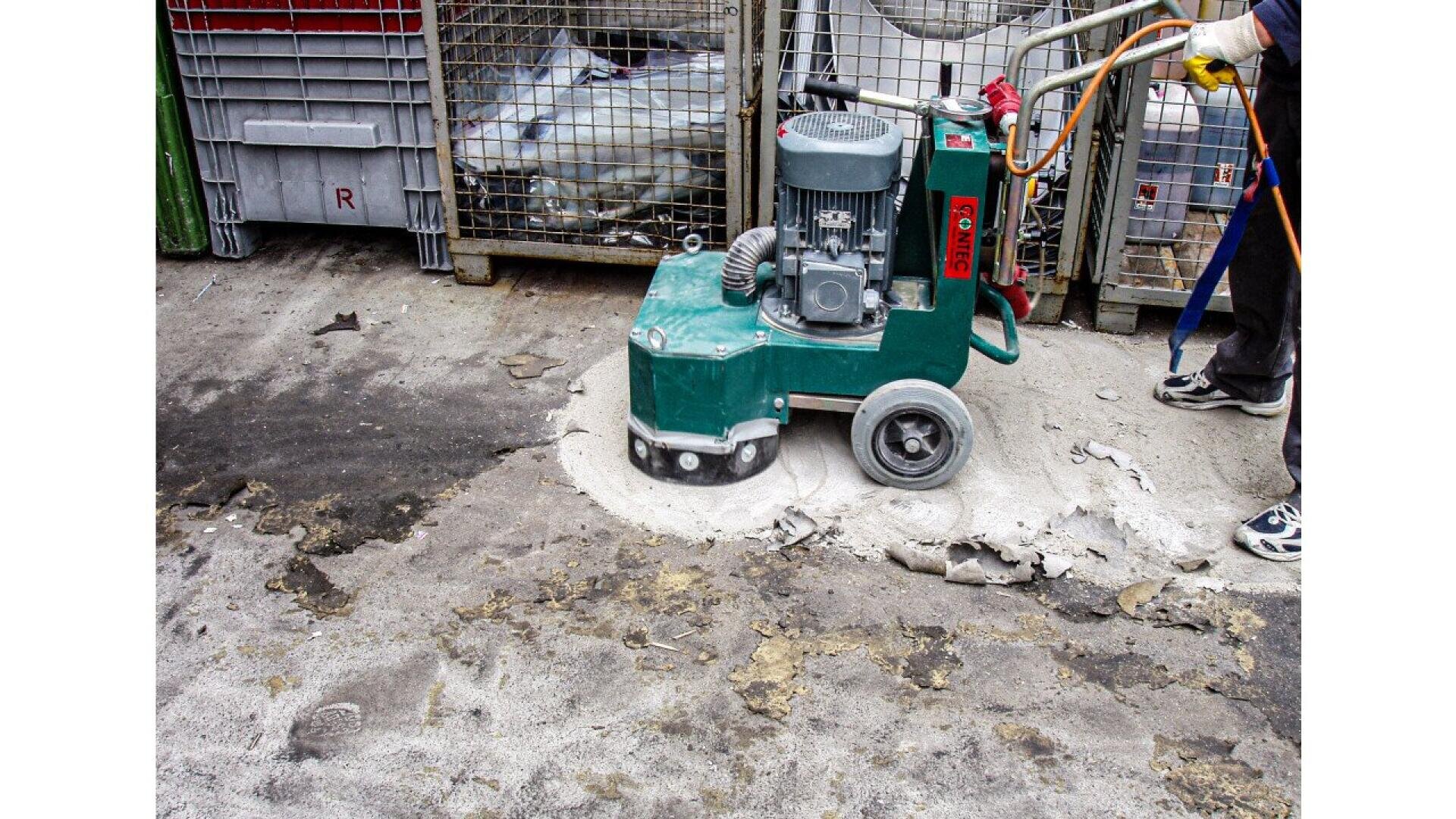 A worker operates a floor grinding machine to remove surface material from a concrete floor in an industrial setting, with storage containers visible in the background.
