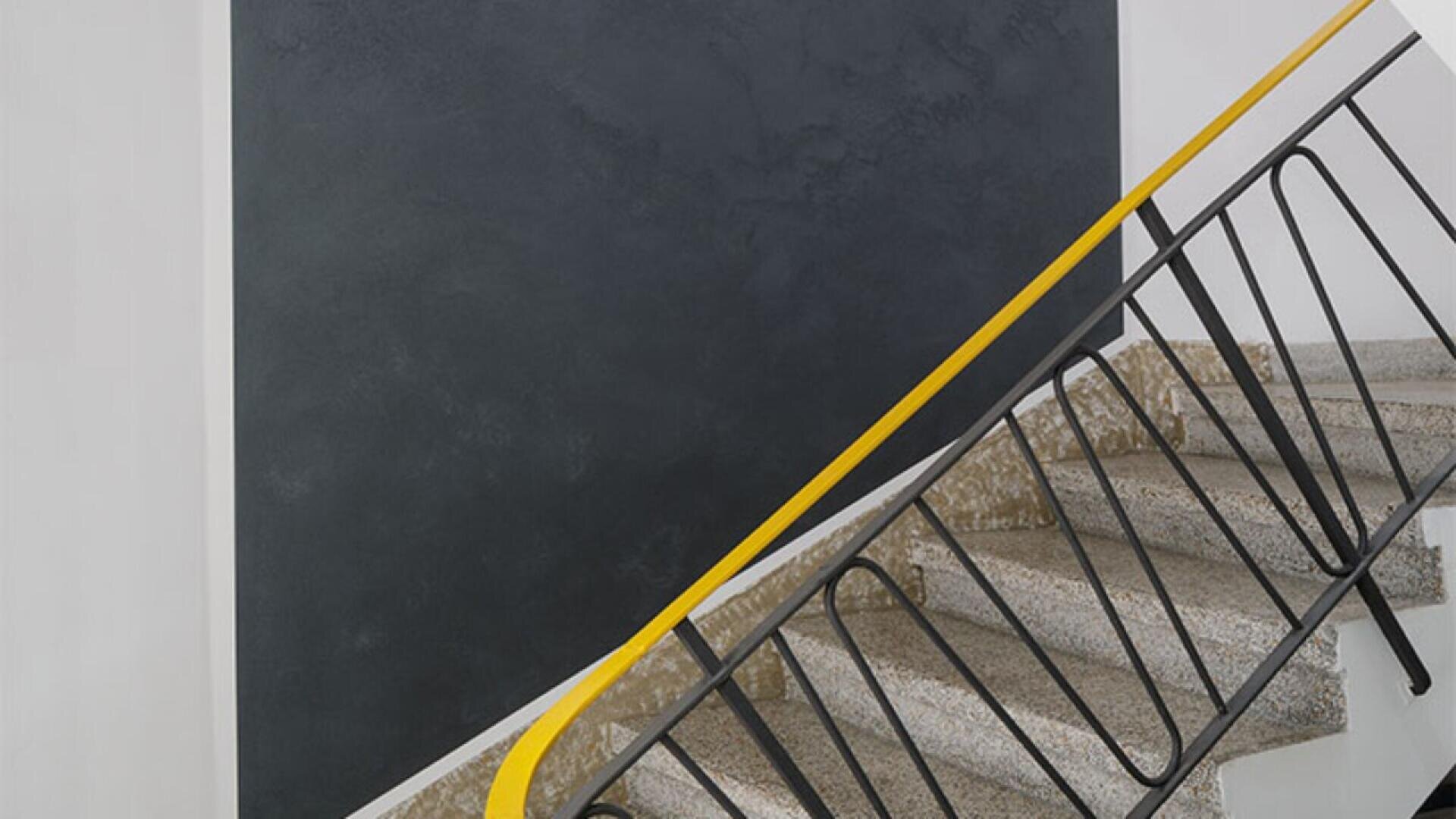 A staircase with terrazzo steps, a black metal railing, and a yellow handrail ascends beside a large, dark gray rectangular wall. The setting has a clean, modern, minimalist design.