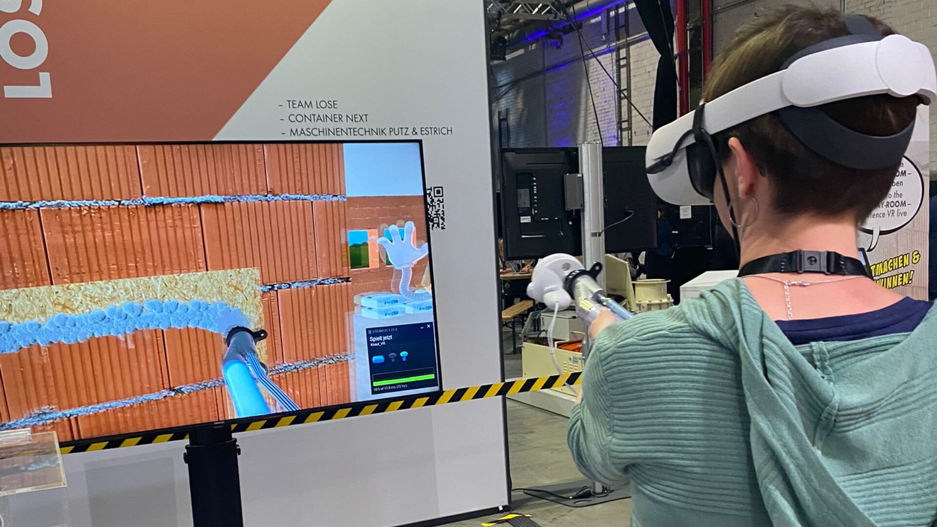 Eine Person mit VR-Headset und Controllern interagiert mit einer Bausimulation auf einem großen Bildschirm. Sie zeigt virtuelles Mauerwerk und Dämmung. In der Nähe sind Industrieanlagen und Absperrbänder zu sehen.