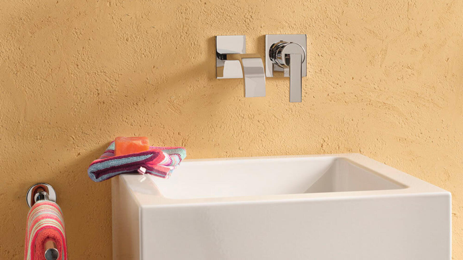 Ein modernes Waschbecken mit einem an der Wand montierten Wasserhahn, ein gestreiftes Handtuch, das an einem Haken hängt, und ein gestreifter Waschlappen mit einem orangefarbenen Stück Seife auf dem Waschbeckenrand, alles vor einer strukturierten beigen Wand.