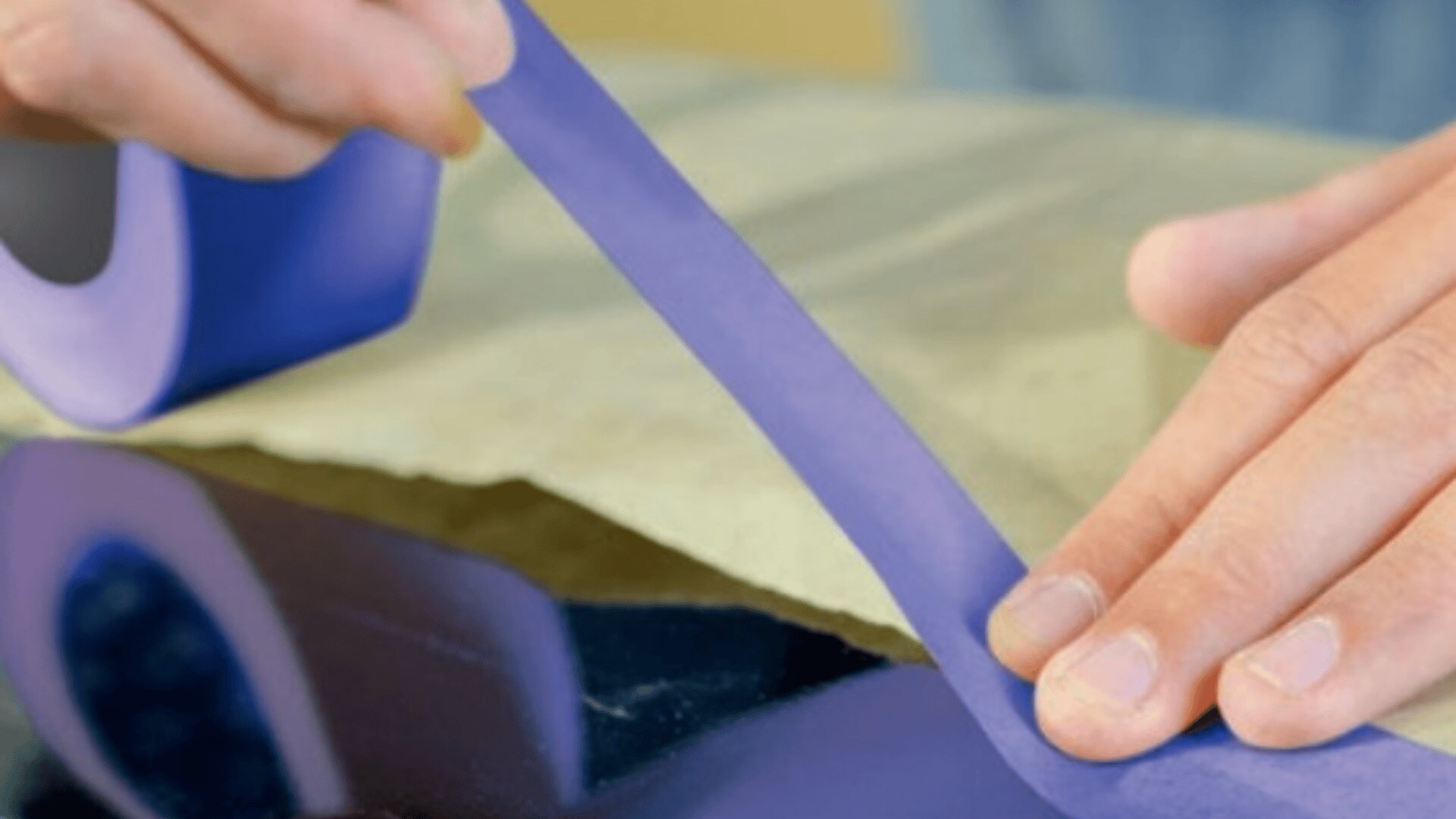 A close-up of hands applying a strip of blue painter’s tape onto a surface, preparing for painting or protection.