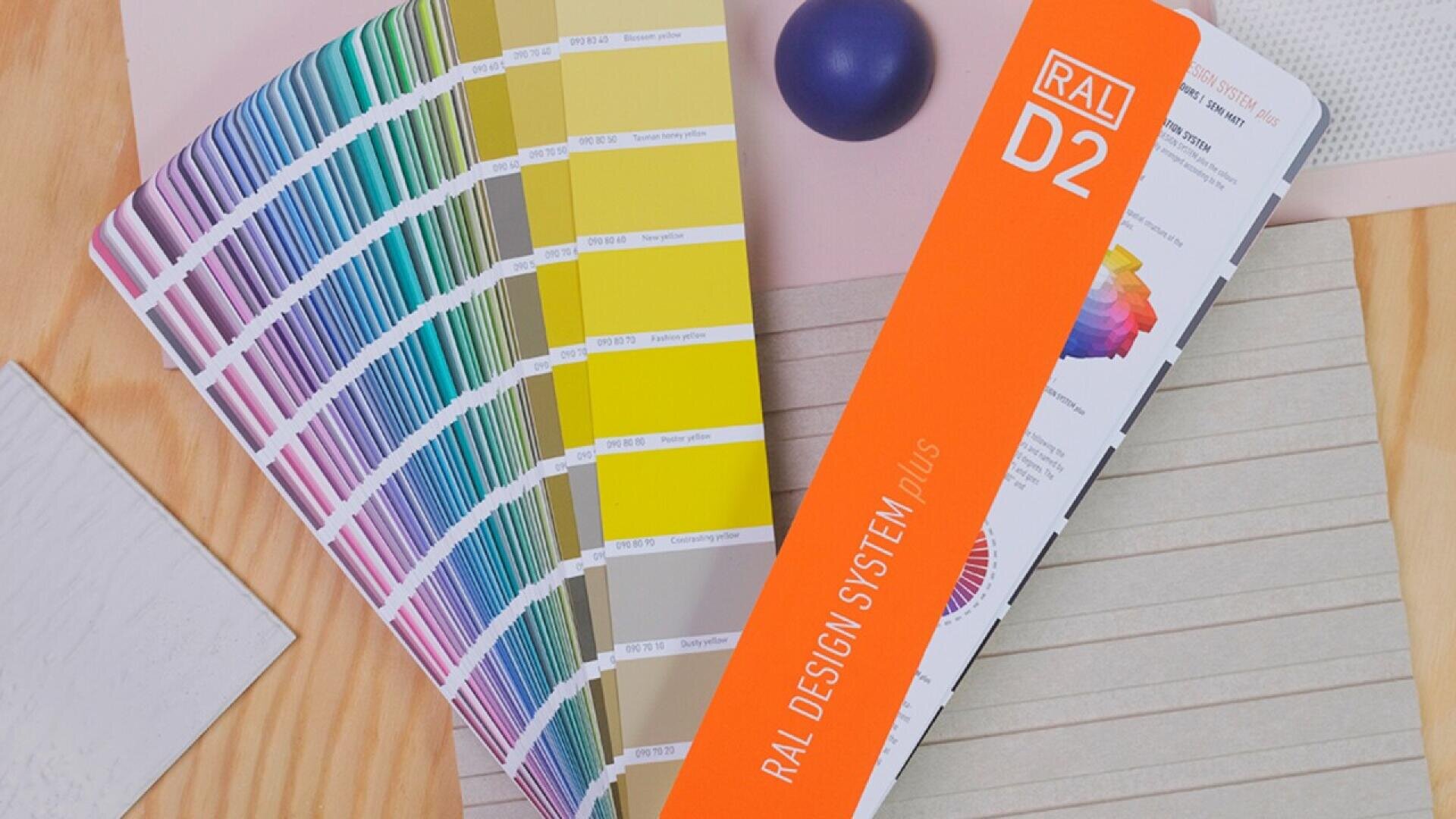 A RAL Design System Plus color fan deck is open on a wooden surface, displaying a wide range of color swatches. There is also a small blue sphere and sheets of textured papers nearby.