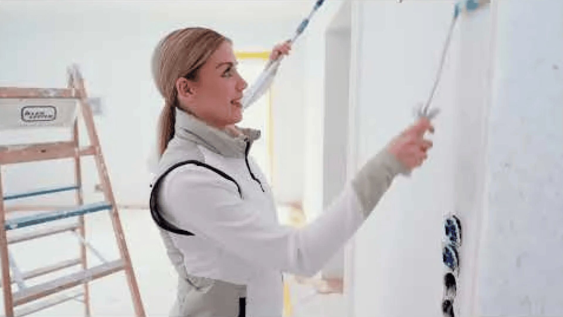 Eine Frau in hellem Outfit streicht mit einer Rolle eine weiße Wand. Im Hintergrund ist in einem hellen, unfertigen Raum eine Leiter zu sehen.