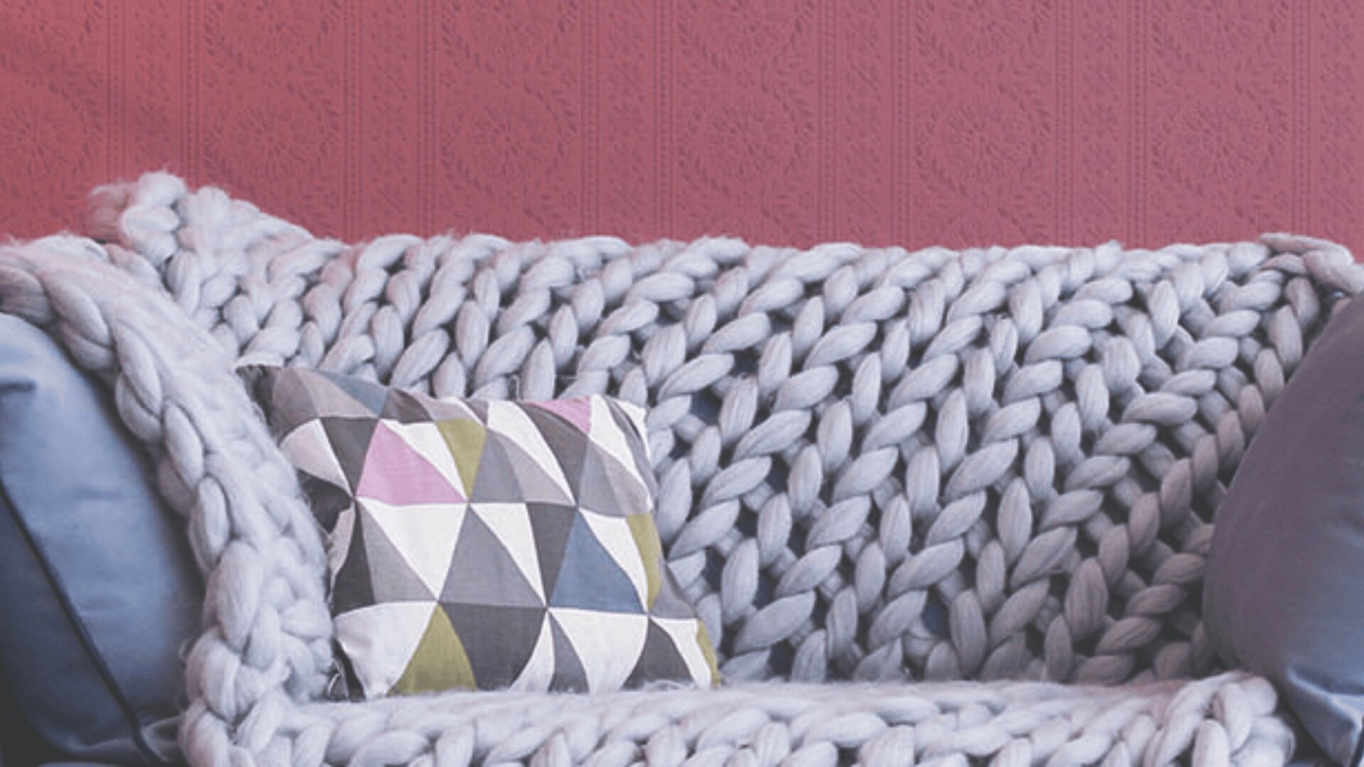 Ein gemütliches Sofa, bedeckt mit einer grob gestrickten Decke und dekoriert mit einem Kissen mit geometrischem Muster, vor dem Hintergrund einer rosafarbenen, strukturierten Tapete.