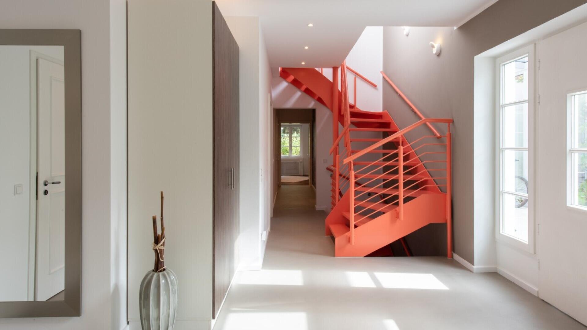A modern hallway with neutral walls and floors features a bold red spiral staircase. Large windows on the right let in natural light. A tall vase and a mirror decorate the space, creating an open, airy feel.