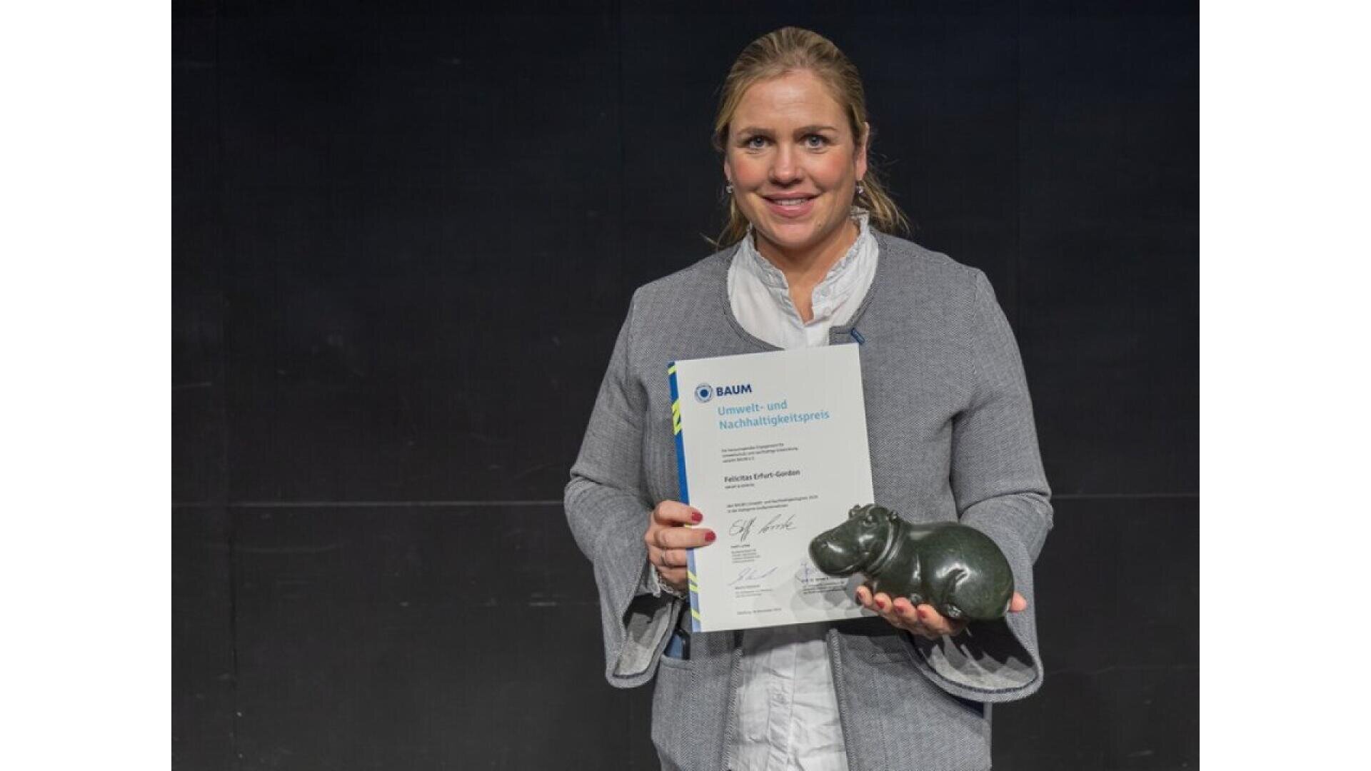 A woman stands against a dark background, smiling and holding a certificate in one hand and a small, dark hippopotamus figurine in the other. She is wearing a gray jacket over a white blouse.