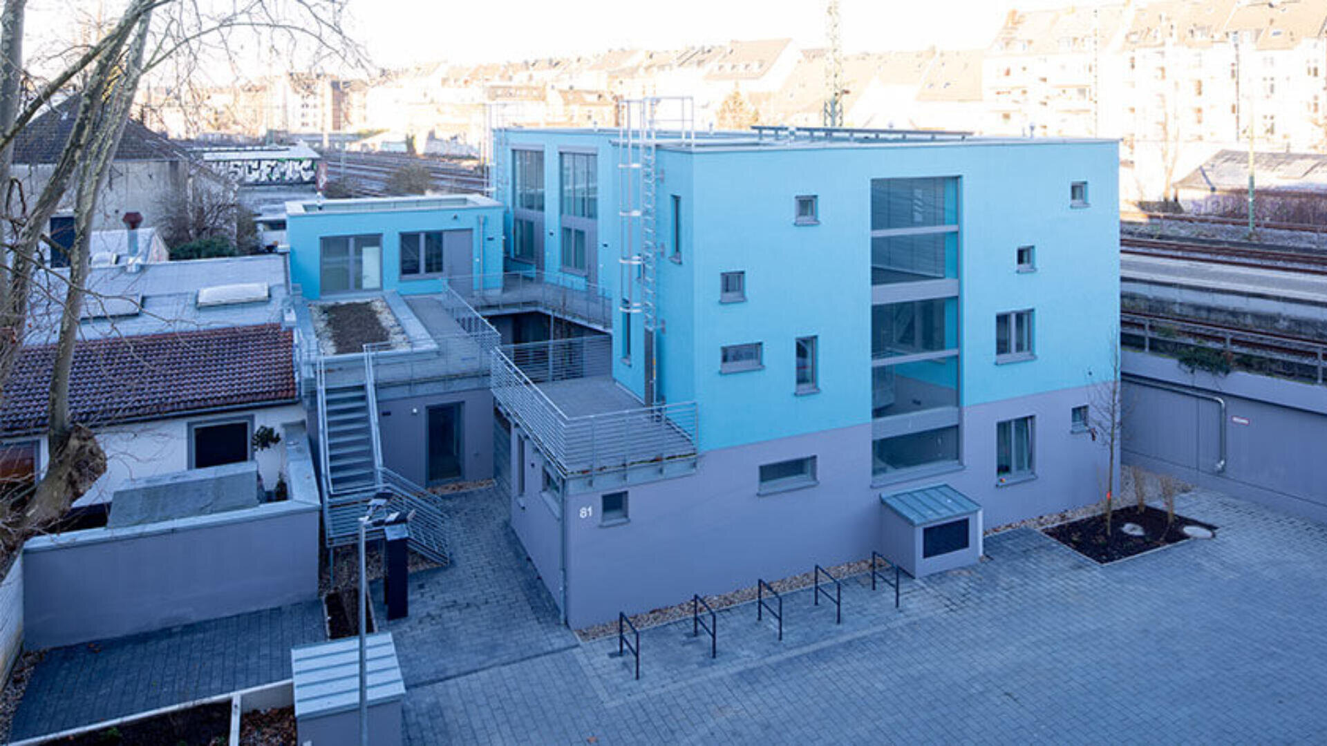 Ein modernes, zweistöckiges, blau-graues Wohnhaus mit Balkonen, Flachdächern und Außentreppen. Der Innenhof verfügt über Fahrradständer und gepflasterte Gehwege. Im Hintergrund sind nahe gelegene Gebäude und Bahngleise zu sehen.