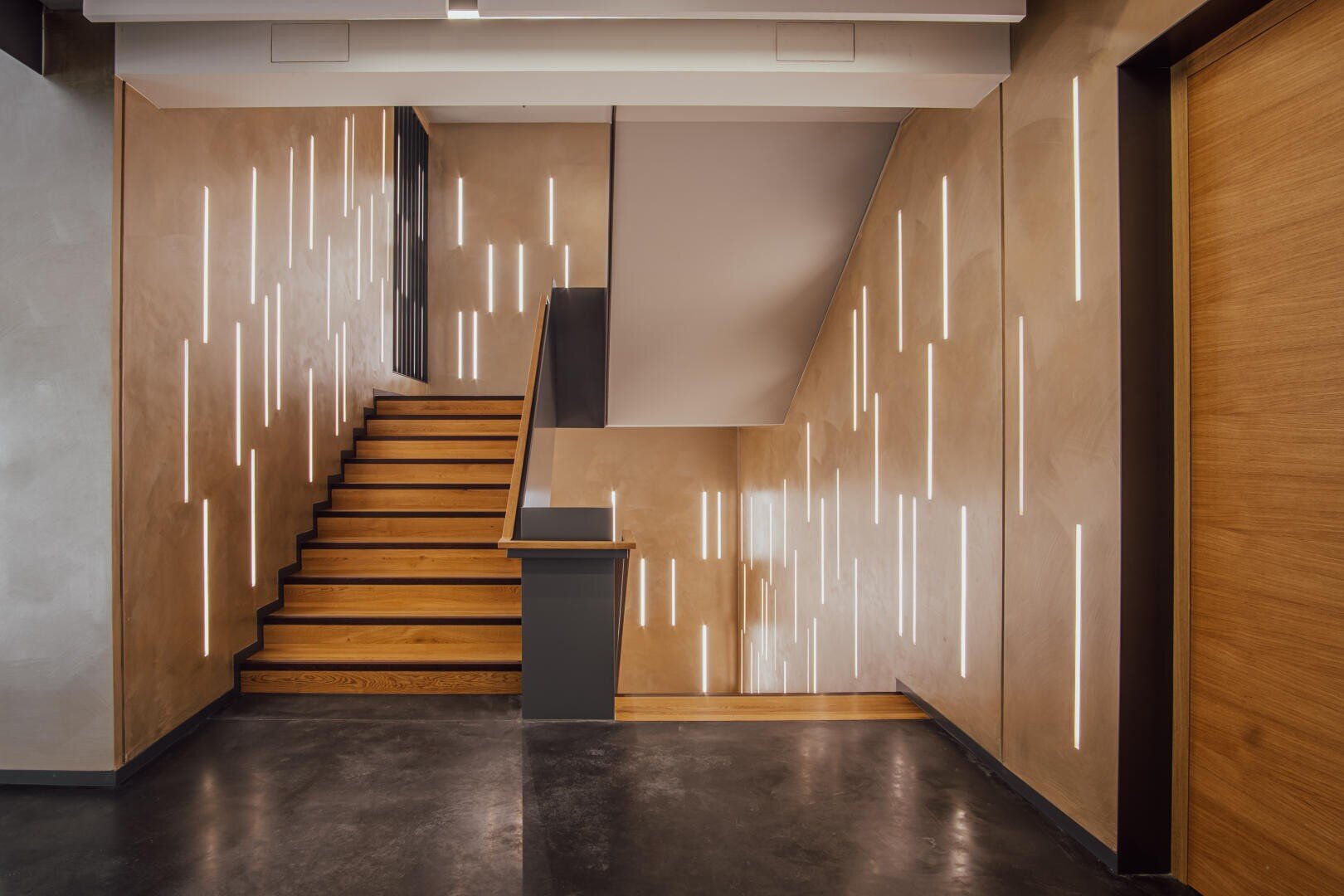Eine moderne Treppe mit Holzstufen und beigen Wänden verfügt über in die Wände eingelassene vertikale LED-Lichtstreifen, die eine stilvolle, zeitgenössische Atmosphäre schaffen.