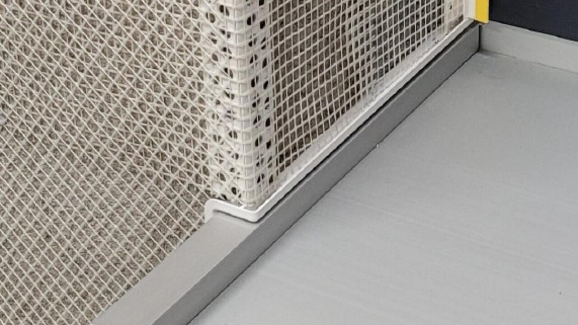 Close-up of a corner where a beige mesh panel meets a light grey floor, held in place by a white L-shaped metal bracket and a small yellow corner guard.