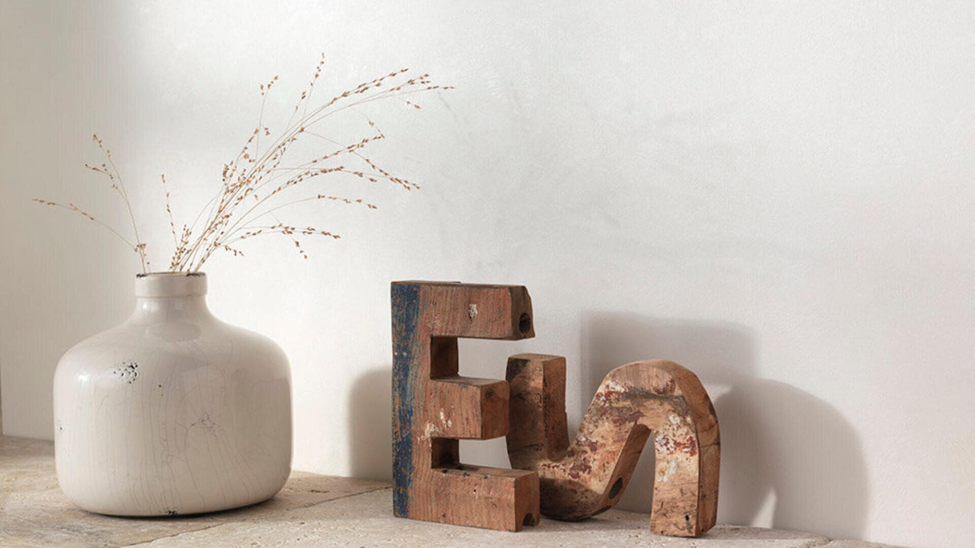 Eine weiße Keramikvase mit getrocknetem Gras steht auf einer hellen Steinoberfläche neben zwei großen, rustikalen Holzbuchstaben, E und N, die an einer schlichten weißen Wand lehnen.