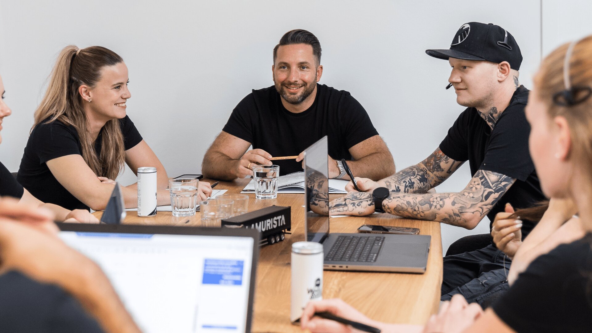 Eine Gruppe von sechs Personen sitzt lächelnd und engagiert um einen Tisch und diskutiert. Laptops, Getränke und Papiere stehen in einem hellen Büro auf dem Tisch.