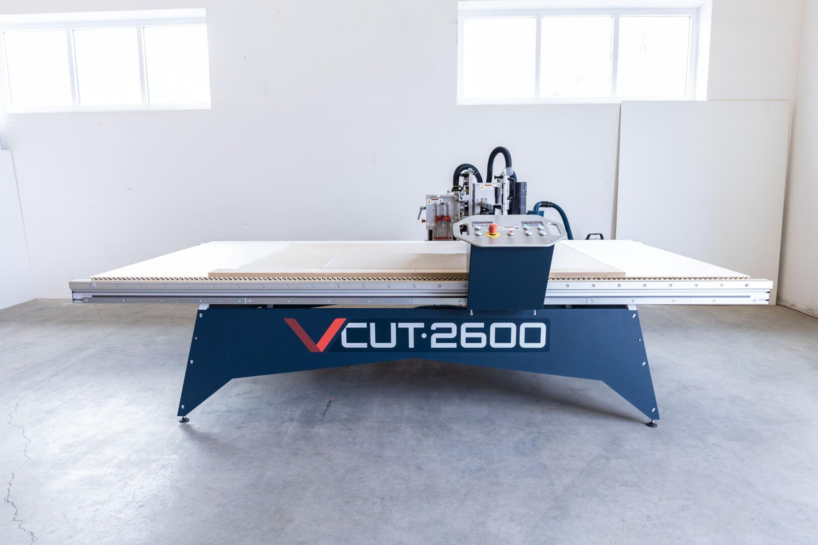 A large industrial CNC machine labeled VCUT 2600 stands in a bright, clean room with white walls and two windows. The machine features a flat working surface and a control panel.