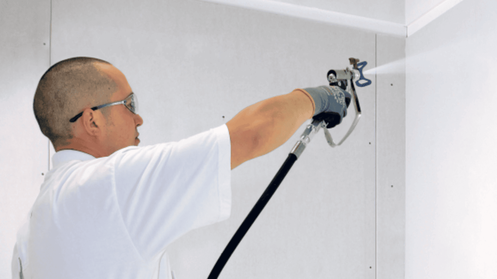 A person wearing safety glasses and gloves uses a spray gun to paint or coat a white wall in an indoor setting.