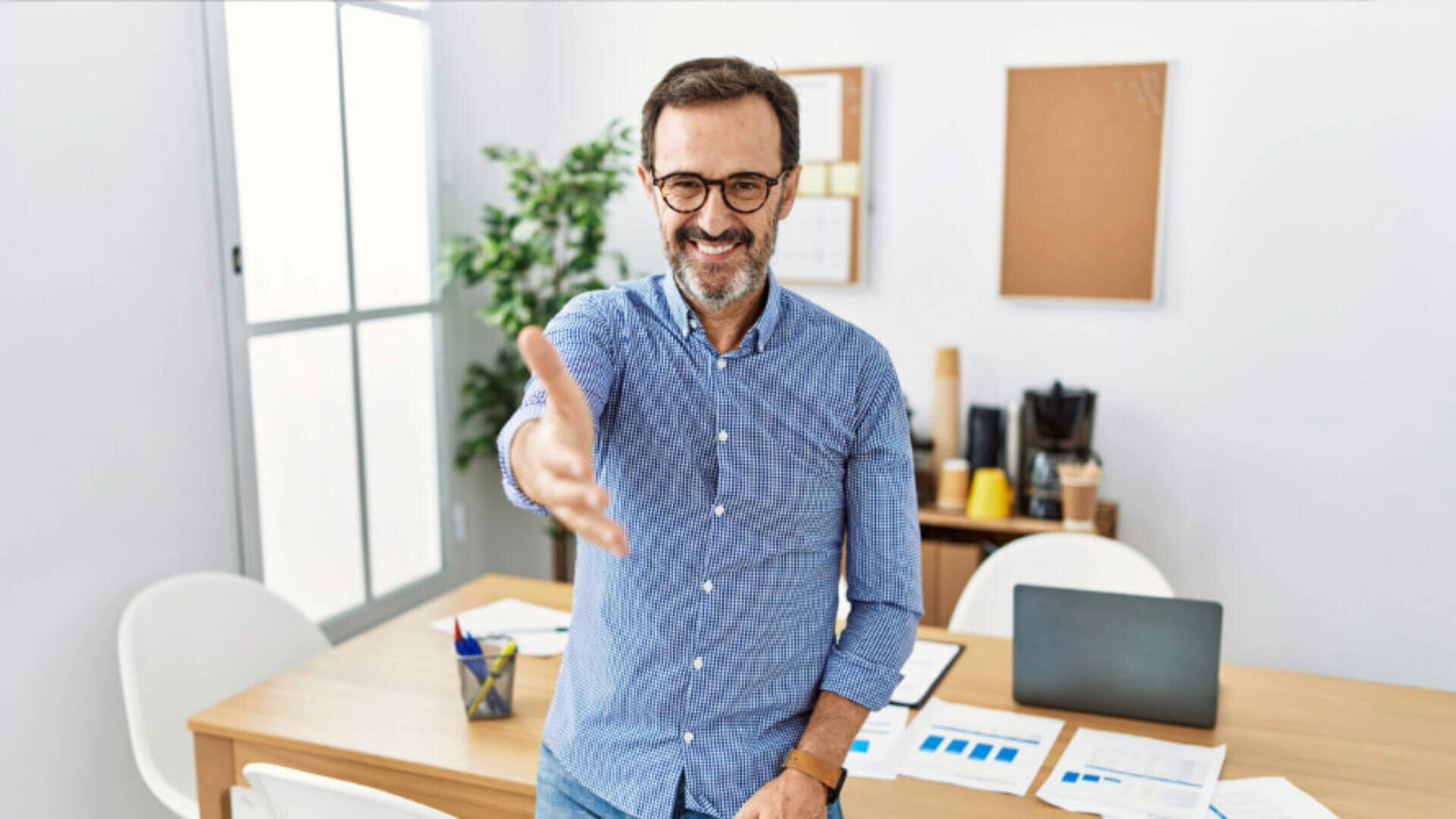 Ein lächelnder Mann mit Brille steht in einem Büro und streckt seine Hand zum Händedruck aus. Auf dem Schreibtisch hinter ihm liegen Papiere, ein Laptop und Büromaterial. Im Hintergrund sind eine Pflanze und Pinnwände zu sehen.