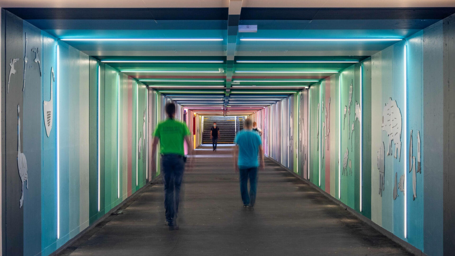 Ein hell erleuchteter Tunnel mit bunten vertikalen Streifen an den Wänden, abstrakten Tierformen und drei Menschen, die hindurchgehen – zwei unscharf im Vordergrund und einer in der Ferne in der Nähe der Treppe.