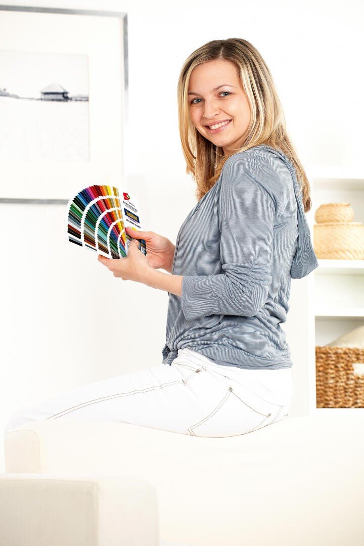 Eine lächelnde Frau mit blondem Haar sitzt seitlich auf einem weißen Sofa und hält einen Farbfächer in der Hand. Sie trägt ein hellgraues Oberteil und eine weiße Hose. Der Hintergrund ist hell mit minimalistischem Dekor und gerahmter Kunst.