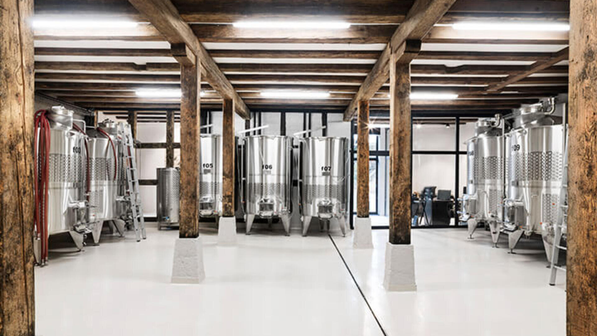 Ein moderner Brauraum mit Gärtanks aus Edelstahl, Holzbalken und weißem Boden. Der Raum ist sauber und gut beleuchtet. In der Nähe der Tanks sind eine Leiter und Geräte sichtbar.