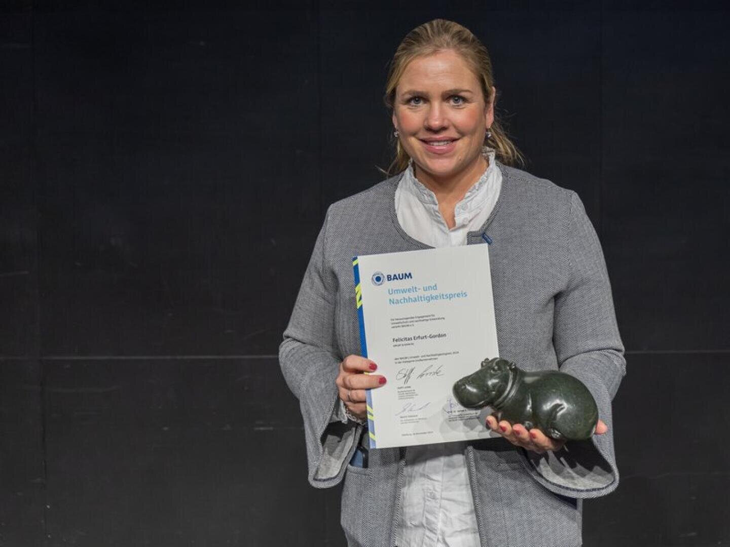 Eine Frau in einer grauen Jacke steht lächelnd vor einem dunklen Hintergrund und hält eine Urkunde und eine kleine Nilpferdskulptur in den Händen, vermutlich als Teil einer Preisverleihung.