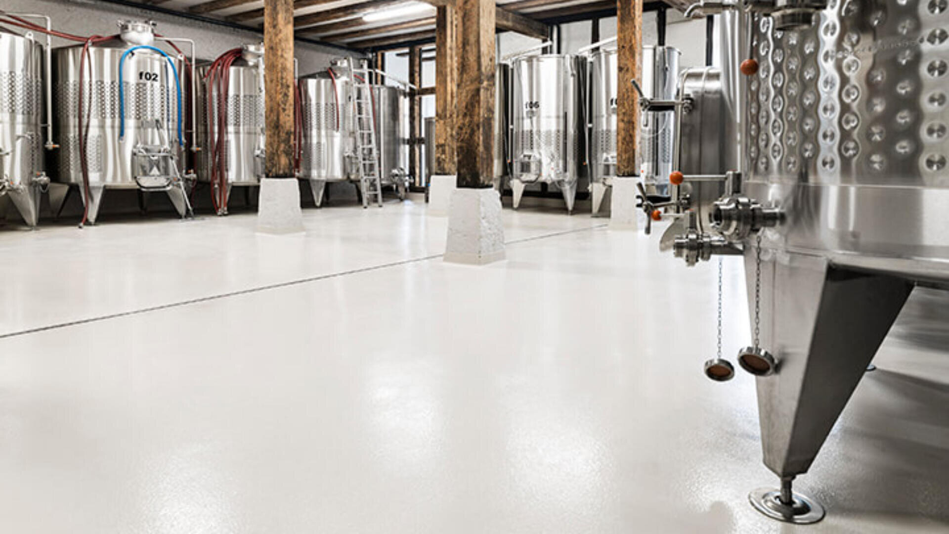 Ein sauberes, modernes Brauerei- oder Weingut-Interieur mit großen Gärtanks aus Edelstahl, hölzernen Deckenbalken und polierten weißen Böden. Im Hintergrund steht eine Metallleiter.